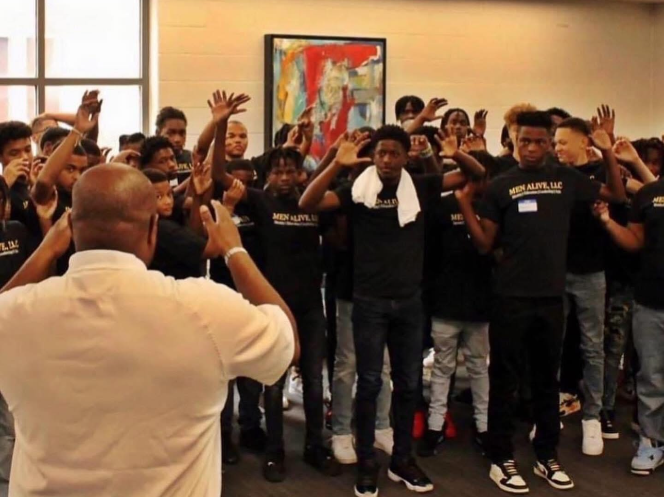 A group of young men participating in a group activity with a leader, some raising their hands, in a room with colorful abstract art on the wall.