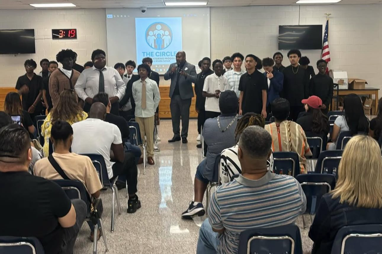 Group of students and a speaker at a middle school presentation titled "The Circle" in a school auditorium with an audience seated and an American flag in the background.