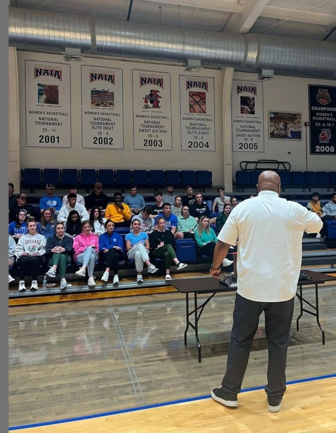 A coach or speaker standing in front of a team of young women sitting on bleachers in a gymnasium. The background shows banners celebrating women's basketball achievements from 2001 to 2006.