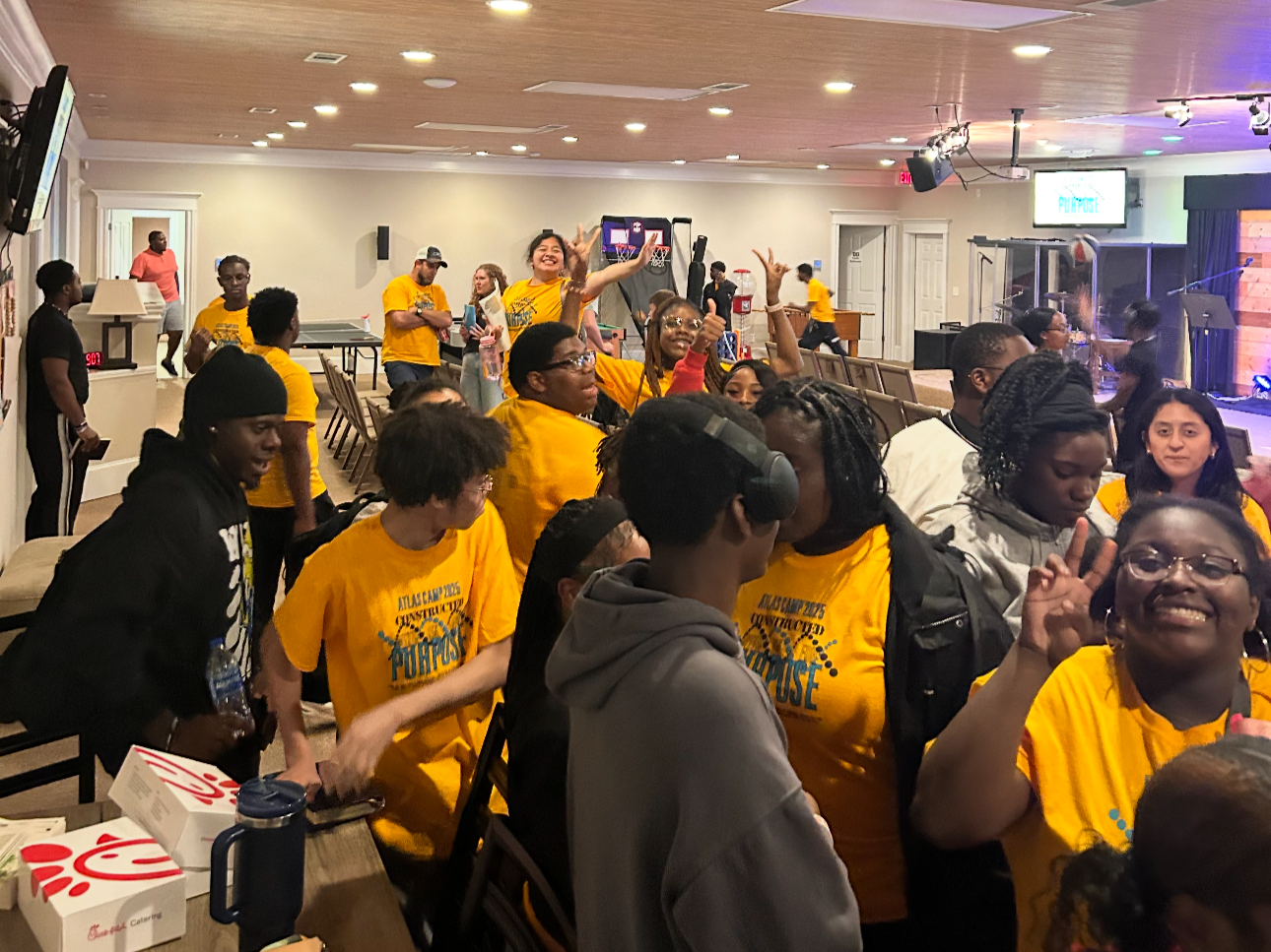 A lively indoor gathering of young people, many wearing yellow T-shirts with Florida Atlantic University and 'Construction Purpose' text, in a decorated event room with stage, screen, and musical equipment, smiling, making peace signs, and mingling.