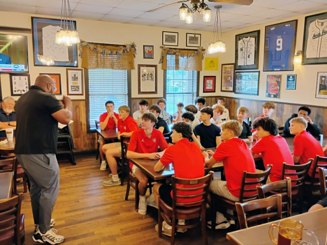A man giving a presentation to a group of young boys in a restaurant decorated with sports memorabilia and framed pictures.