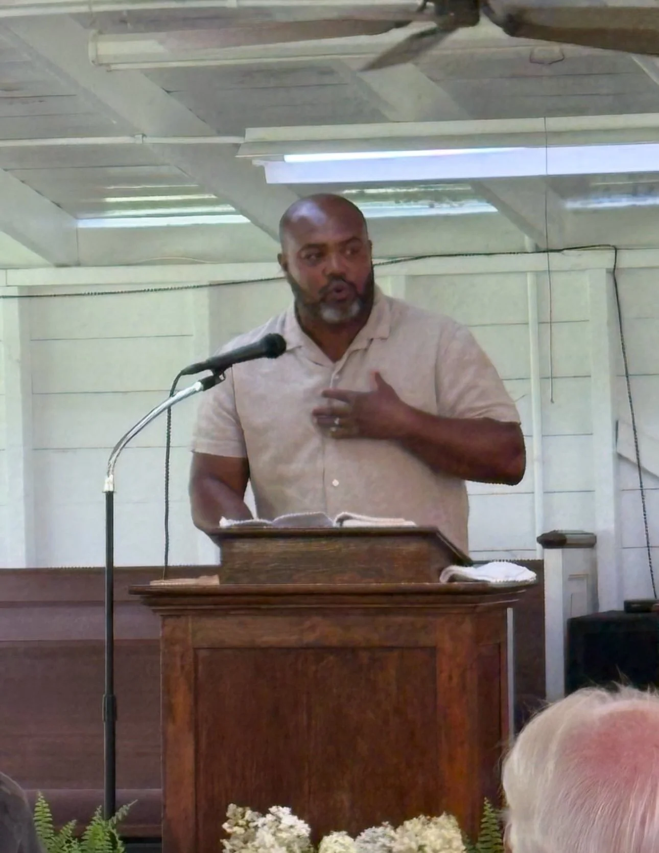 A man with a shaved head and beard speaking at a wooden pulpit, gesturing with his left hand over his chest, in front of a microphone and open Bible, in a room with light-colored wooden walls and ceiling.