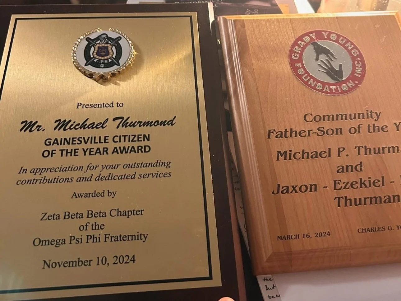 Two wooden plaques, one gold and one brown. The gold plaque has an insignia at the top with crossed swords, a shield, and a ribbon, and it is awarded to Mr. Michael Thurmond for the Gainesville Citizen of the Year Award, presented by the Zeta Beta Be