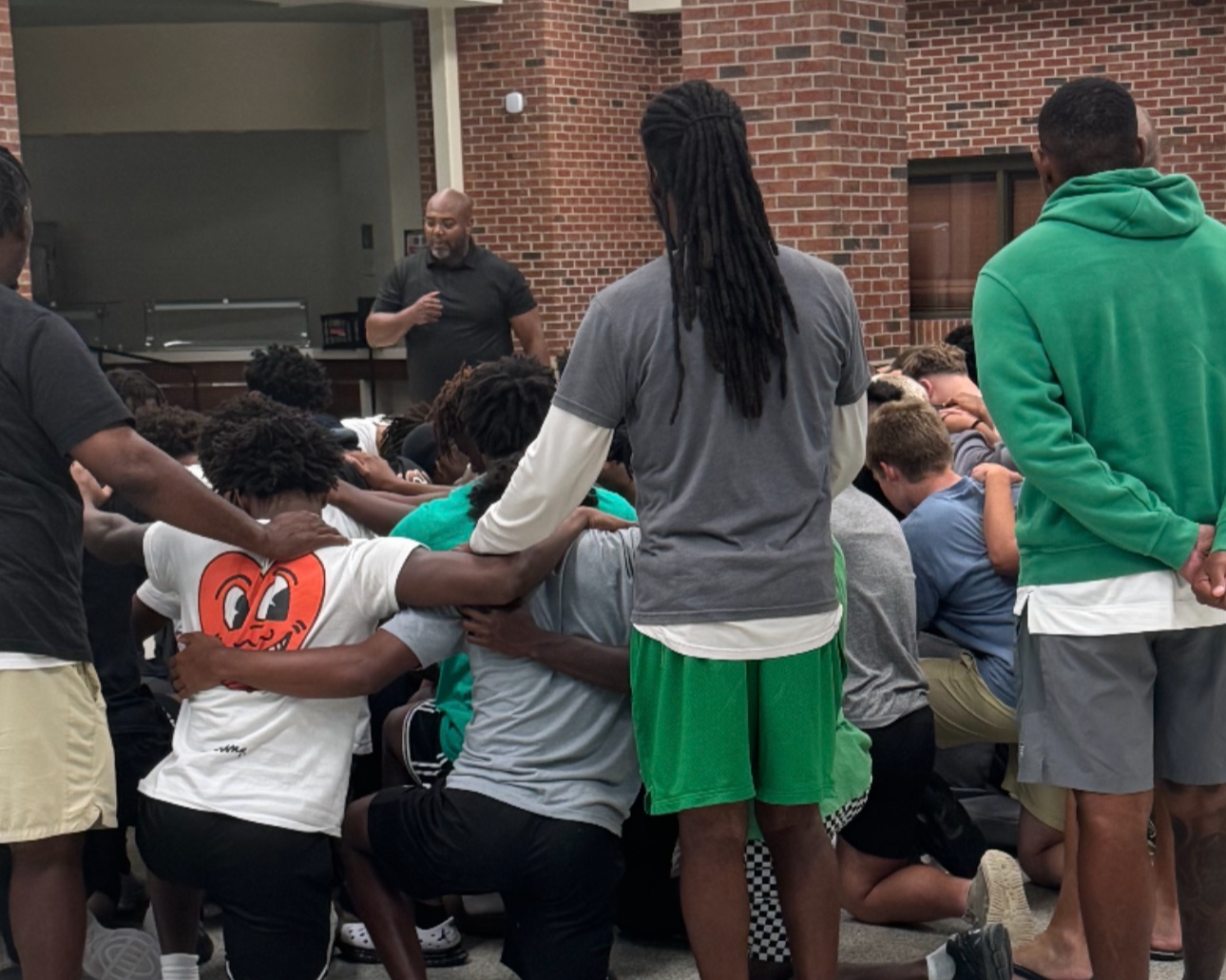 Group of young men participating in a prayer circle with their heads bowed, some with hands on others' shoulders, led by a man standing in front, inside a room with brick walls.