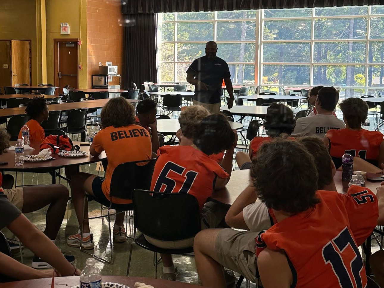 A group of young football players sitting around tables listening to a man giving a presentation in a spacious room with large windows and natural light.