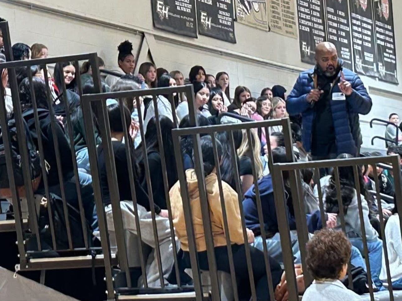 A group of students seated on bleachers in a gymnasium, listening to a man speaking. There are banners on the wall behind them.