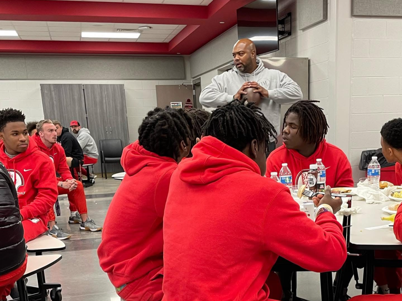 A group of young men, some wearing red hoodies, sitting at a cafeteria table with food and drinks, listening to a man standing and holding a football in a cafeteria or gymnasium setting.