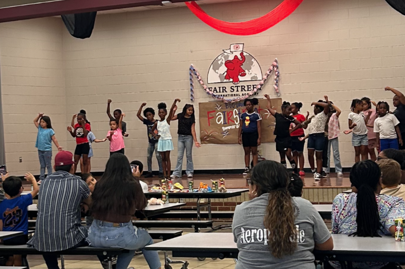 Children performing on stage at a school event, with an audience of adults and kids watching.