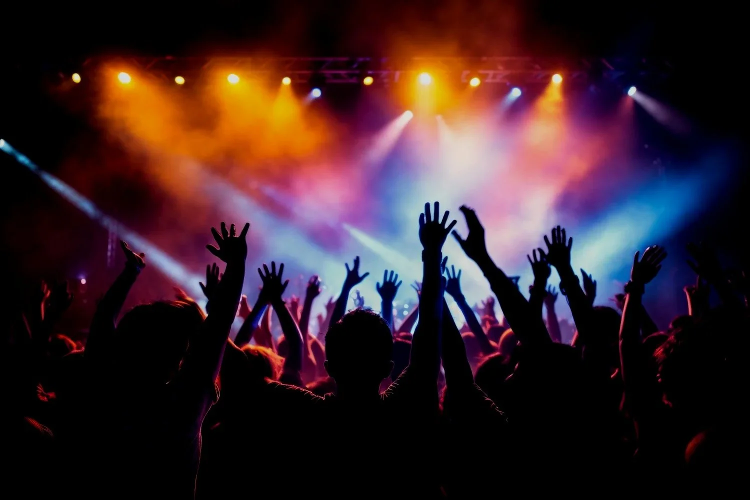 Crowd of people with raised hands at a concert with colorful stage lights.