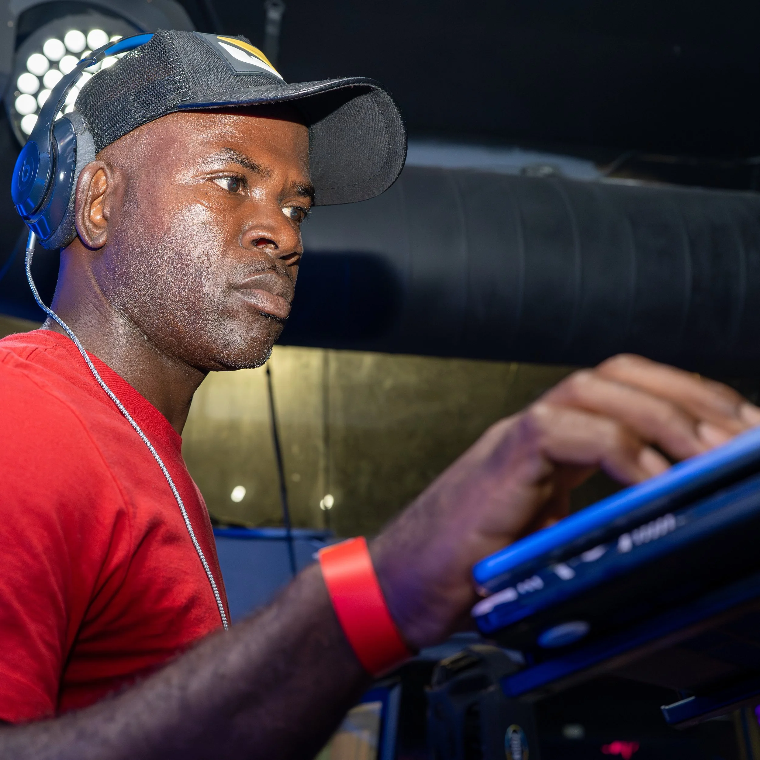 A young man wearing a black baseball cap with a mesh back, large over-ear headphones, and a red shirt, looking at a smartphone. He is sitting in a vehicle, likely a bus or train, with bright lights visible in the background.