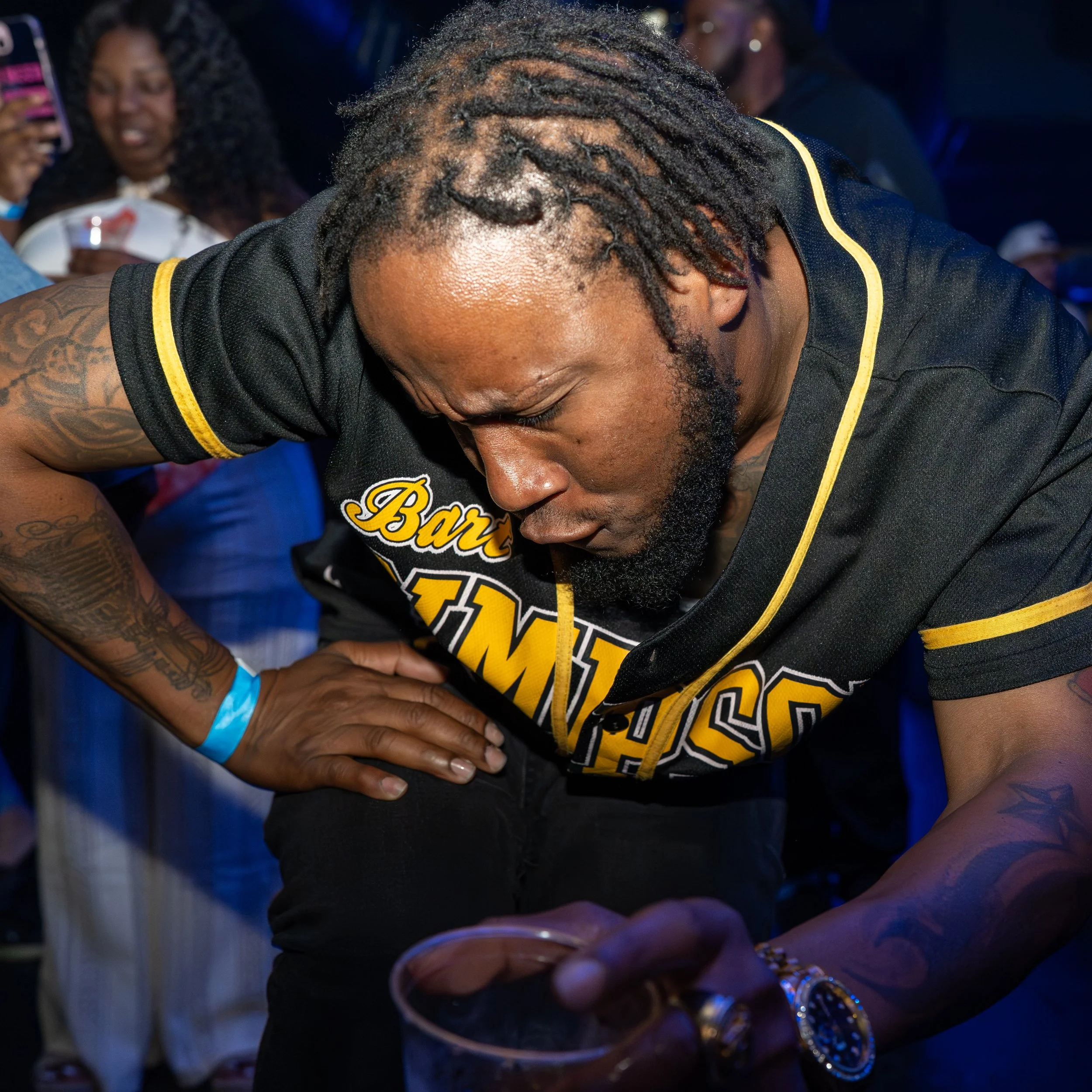 Close-up of a man with dreadlocks and tattoos leaning over a table with a drink, surrounded by people in a party setting with blue and purple lighting.