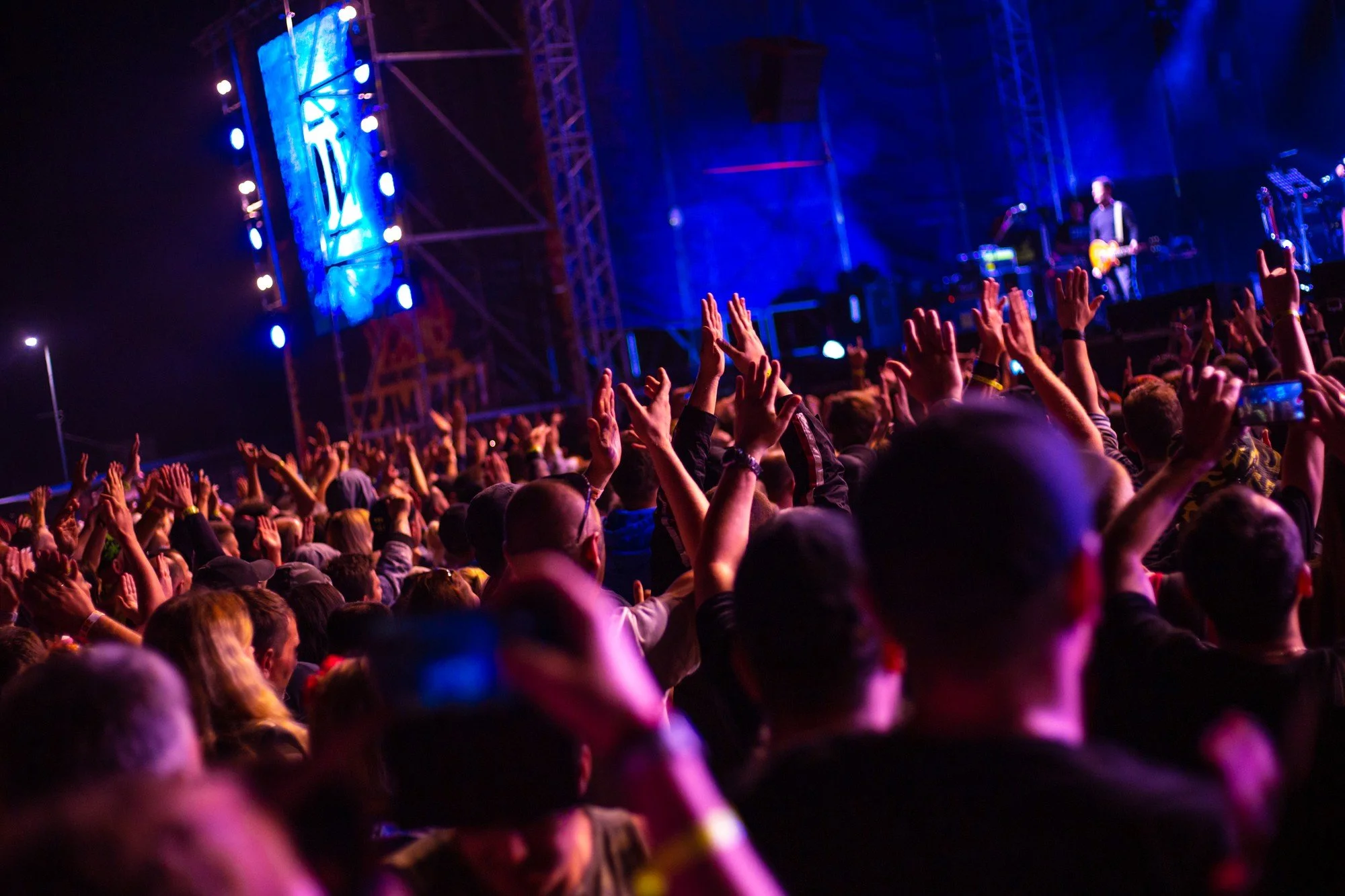 A large crowd of people at a concert with their hands raised, facing a stage with a performer playing guitar, illuminated by blue and purple lights at night.