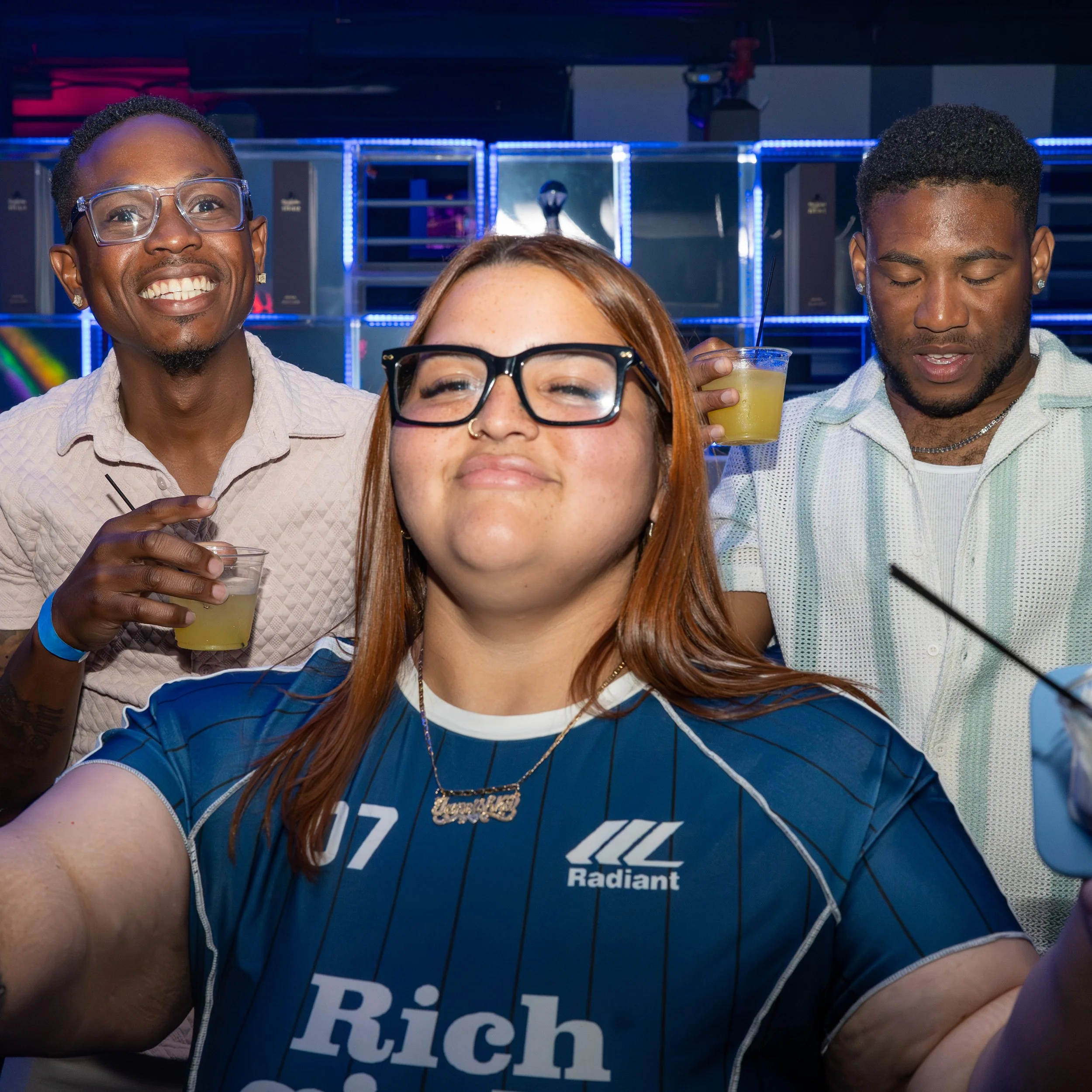 Four people at a social gathering, two men and two women, with a background of neon lights and shelves with various items. The man on the left is smiling, wearing glasses, and a white shirt, holding a drink. The woman in the front center has red hair
