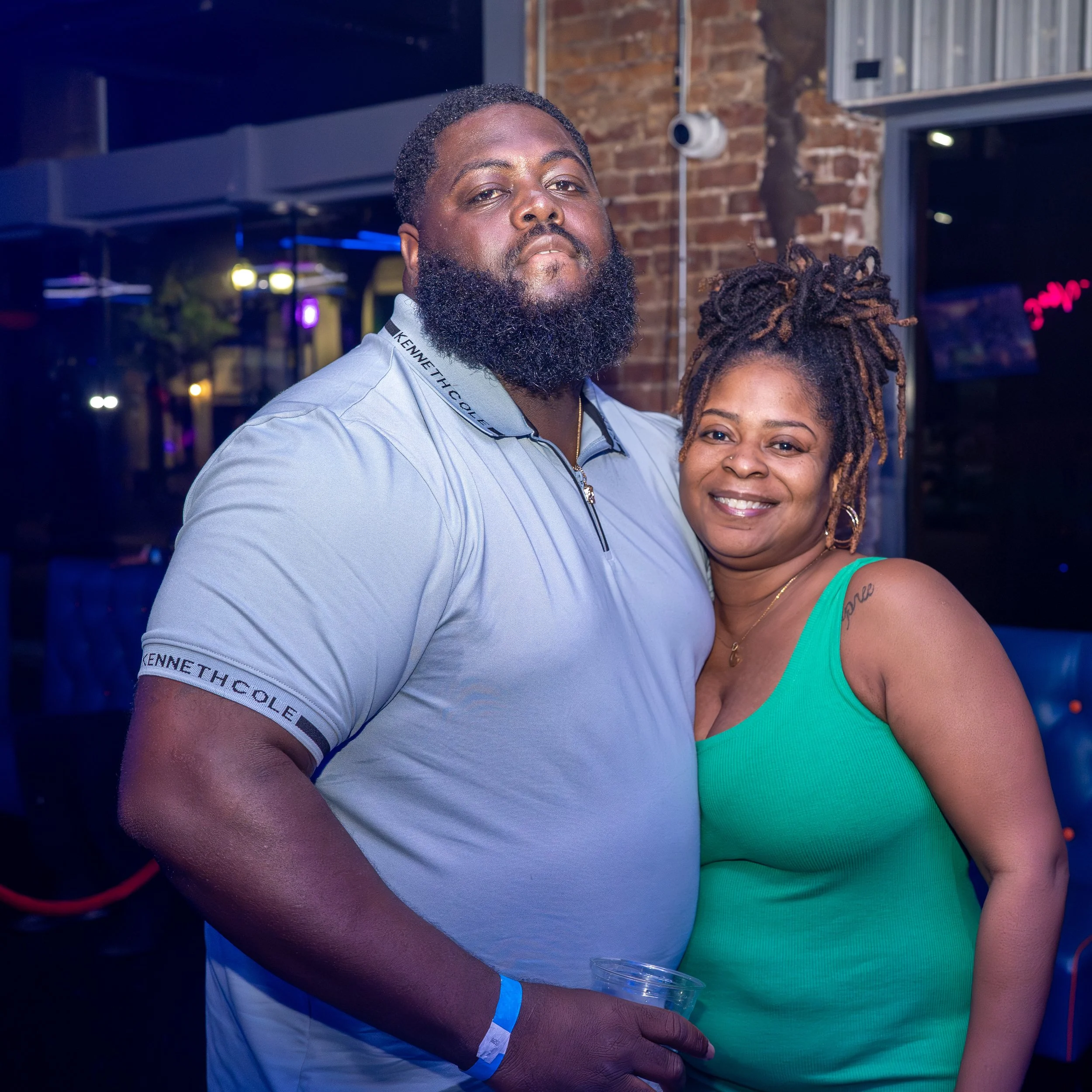 A man with a beard and short hair standing close to a woman with dreadlocks, both smiling at a party or social gathering with brick walls and neon lights in the background.
