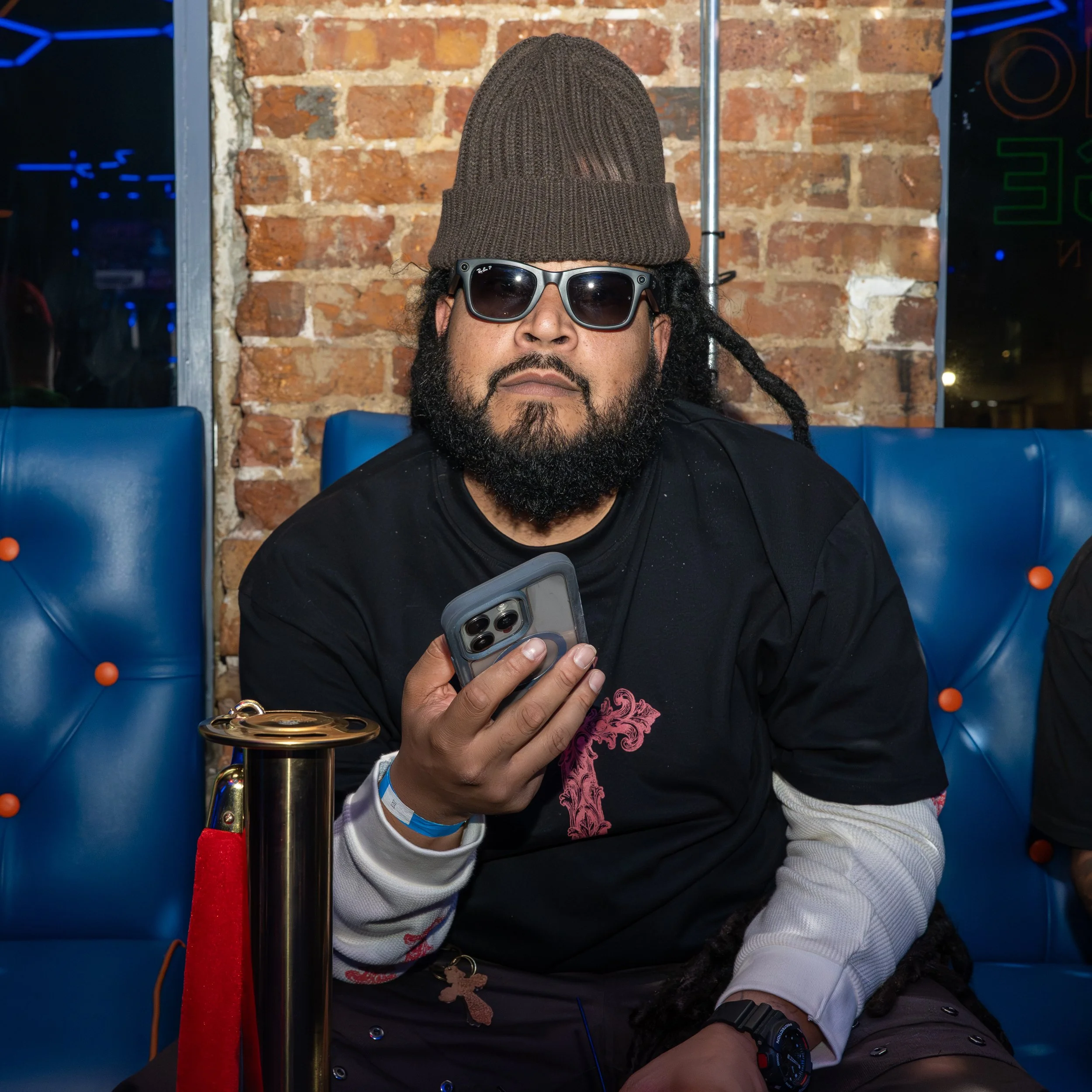 A man with a beard wearing sunglasses, a brown beanie, a black shirt with pink embroidery, and a wristwatch, sitting against a brick wall, holding a smartphone in his hand.