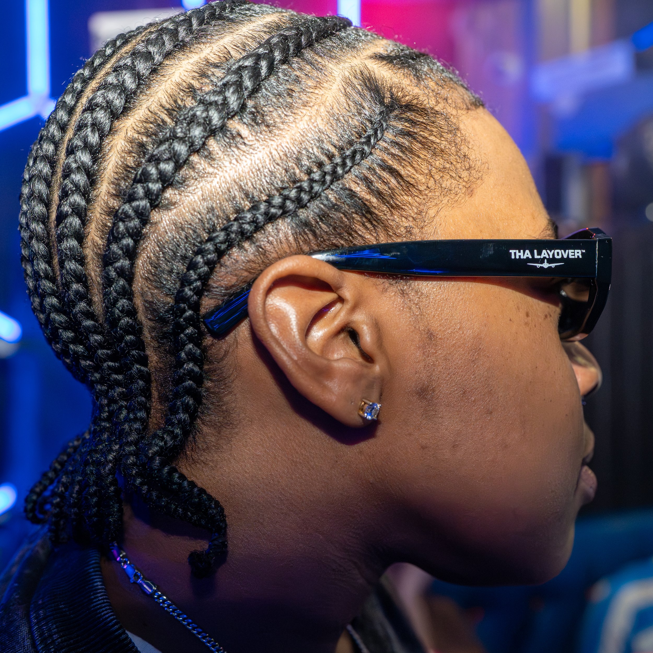 Close-up of a person's head with neatly styled braids, wearing blue-tinted sunglasses with 'THA LAYOVER' written on the side, and a dark, textured shirt against a blurred colorful background.
