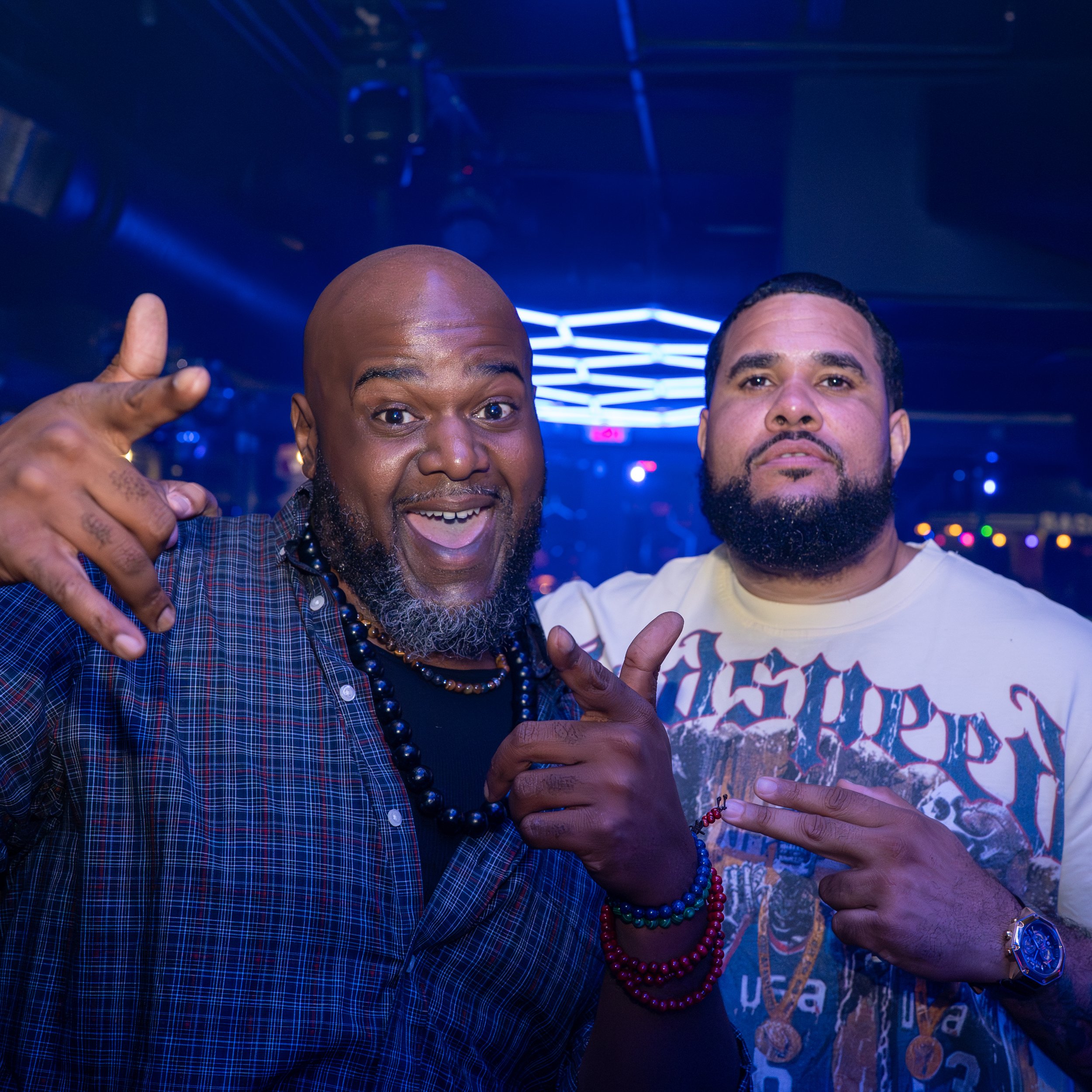 Two men smiling and posing for a photo in a dimly lit room with blue neon lights. One man has a bald head and a beard, wearing a plaid shirt with beaded necklaces. The other man has dark hair and a mustache, wearing a T-shirt with graphic print. Both
