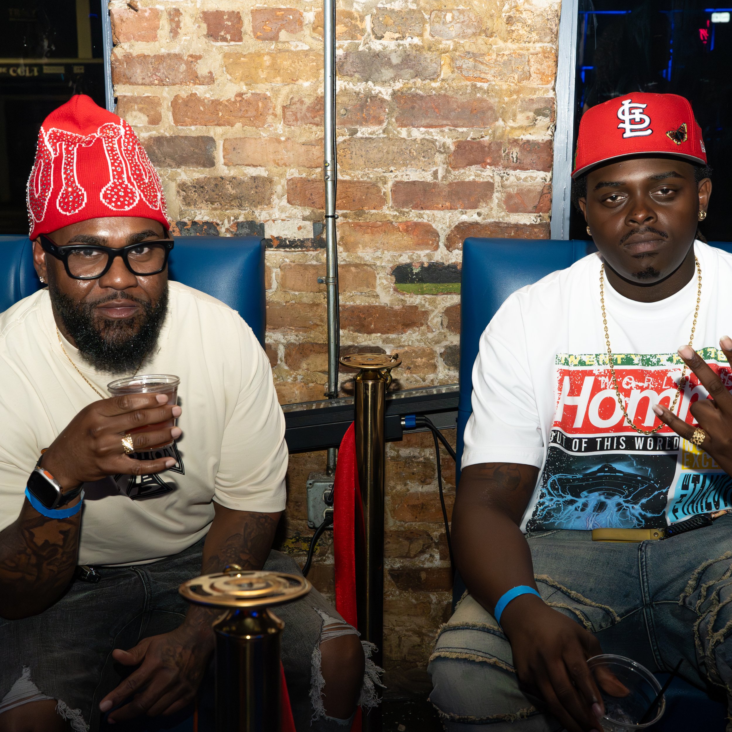 Two men sitting side by side in front of a brick wall, wearing red hats, one with glasses and the other with jewelry, holding drinks, partially visible in the image.