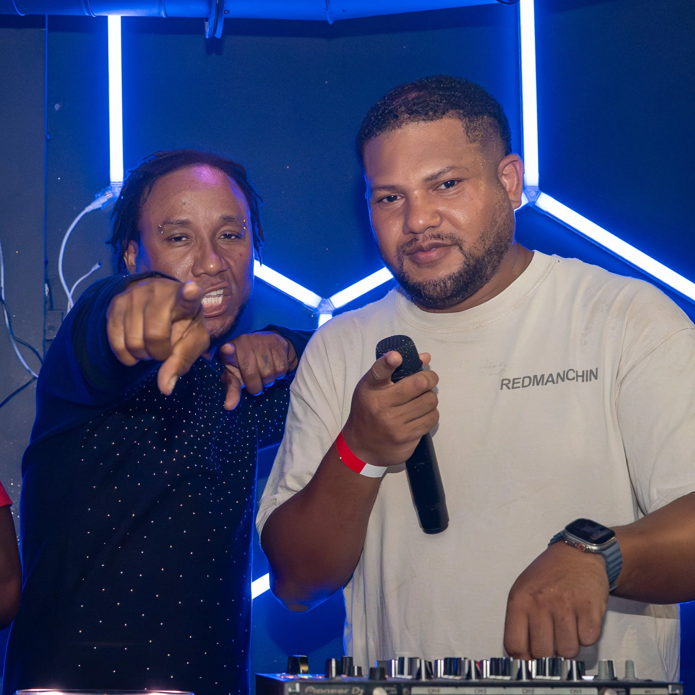 Two men are posing for a selfie. One is holding a microphone, and the other is pointing at the camera. They are standing in a room with blue neon lights and a dark background.