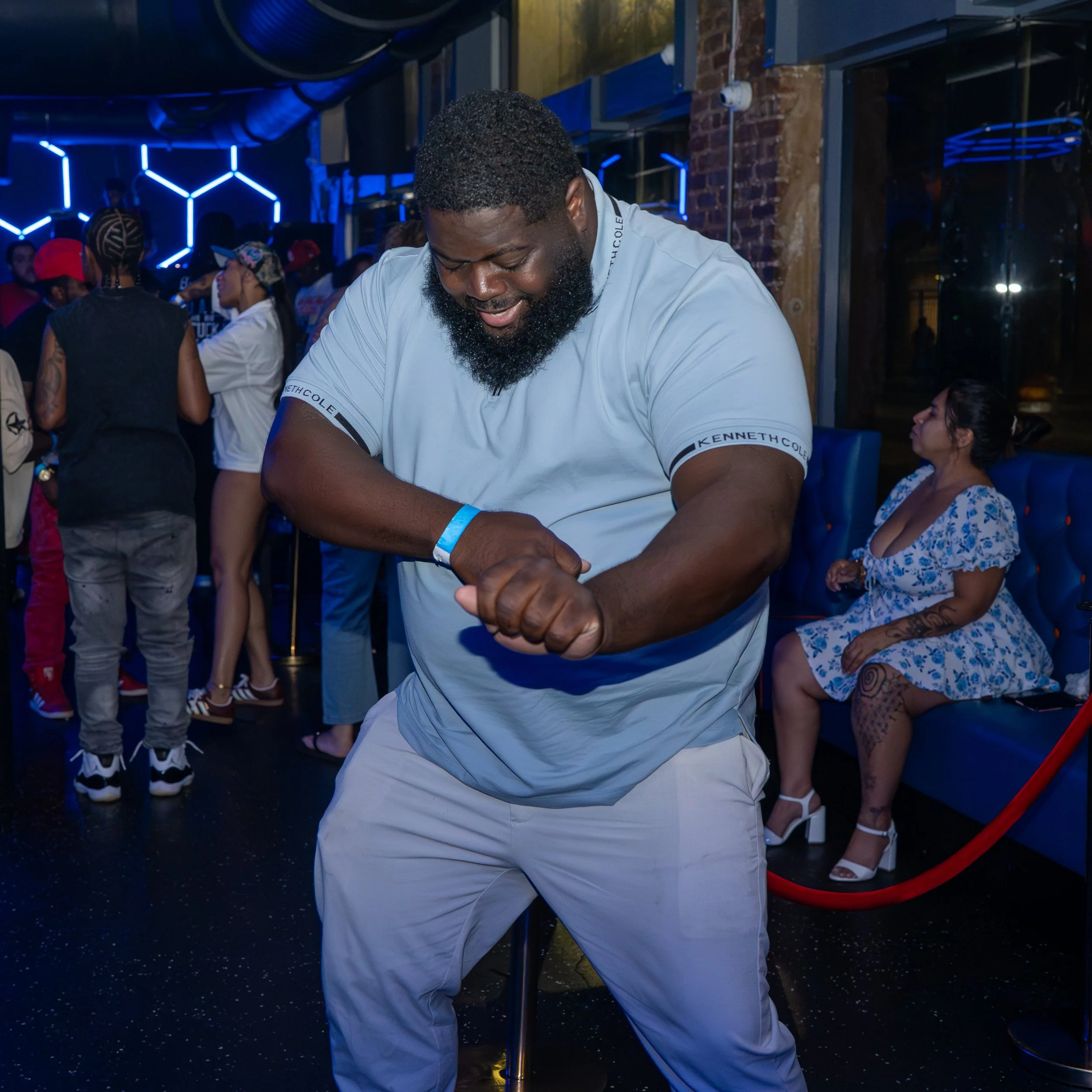 A man with a beard and curly hair wearing a white T-shirt with black text is dancing at a party or nightclub. He appears to be enjoying himself as he dances with his eyes closed and arms raised. The background shows other people socializing, colorful