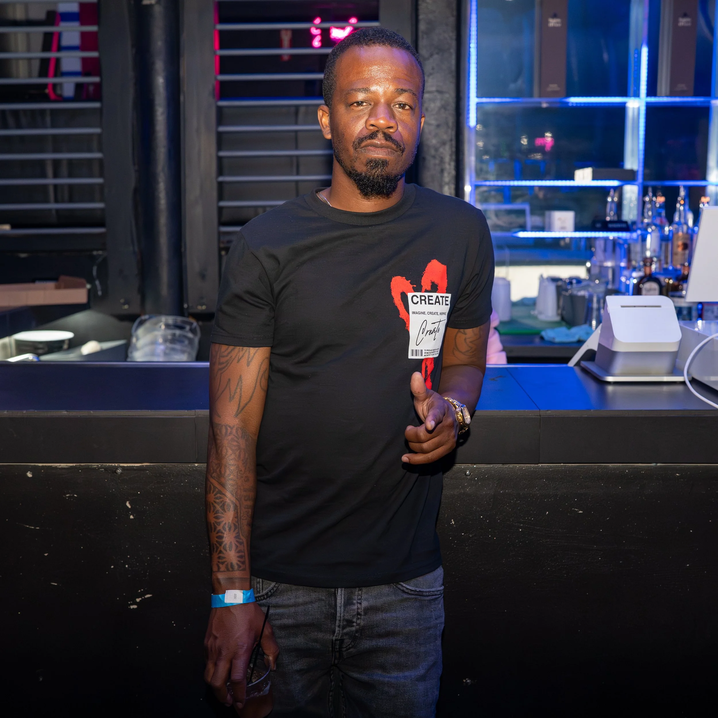 A man with a beard and tattoos on his arm, wearing a black t-shirt with a red heart design and the word 'CREATE' on it, stands in a bar or club setting with dark walls, bright neon lights, and shelves with bottles and glasses in the background. He is