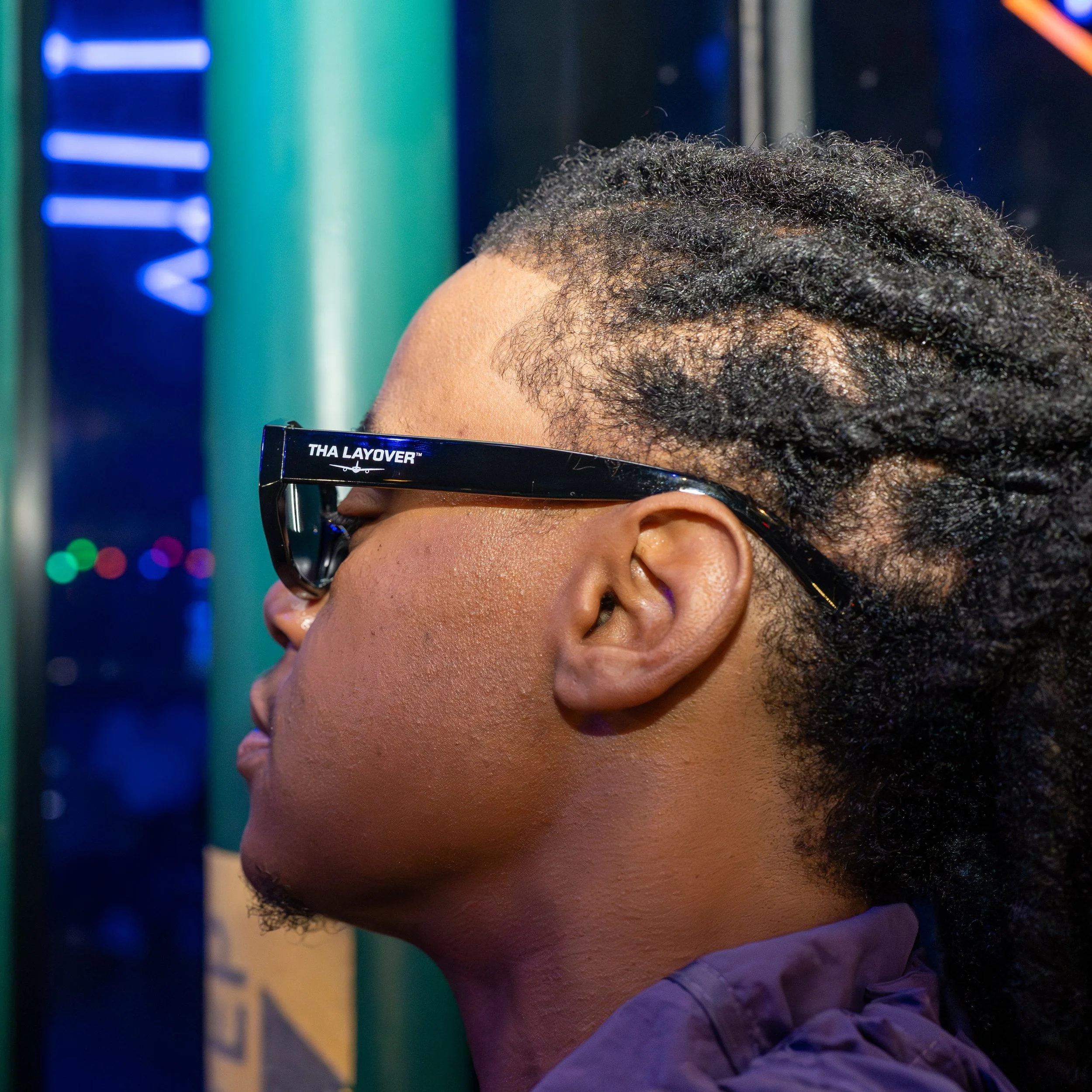 A person with curly black hair wearing black glasses and a white shirt, seen from the side with their face slightly turned down. The background includes colorful lights and some visible text.