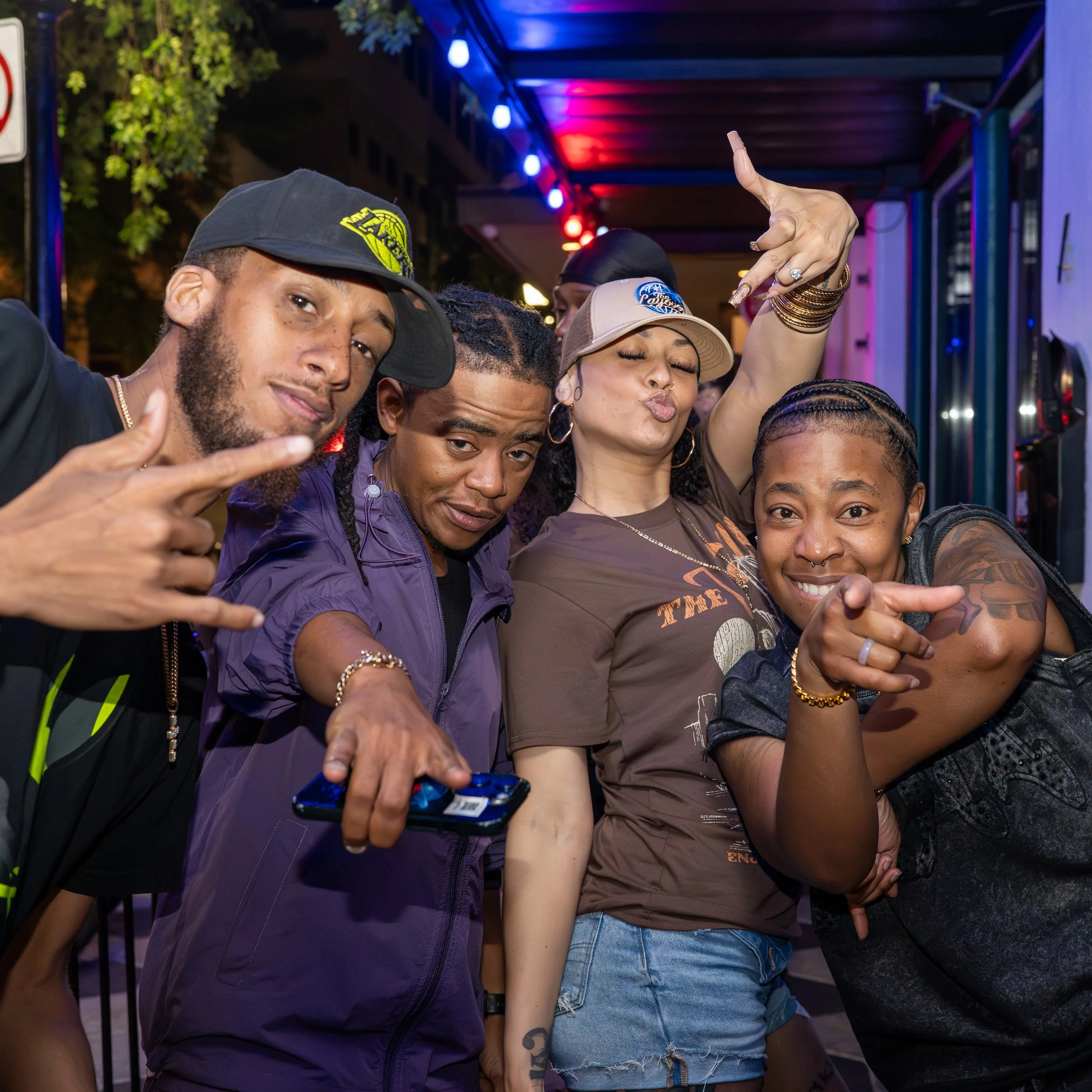 A group of people posing closely together during a night event, some making hand gestures, under colorful lights, with trees and building parts in the background.