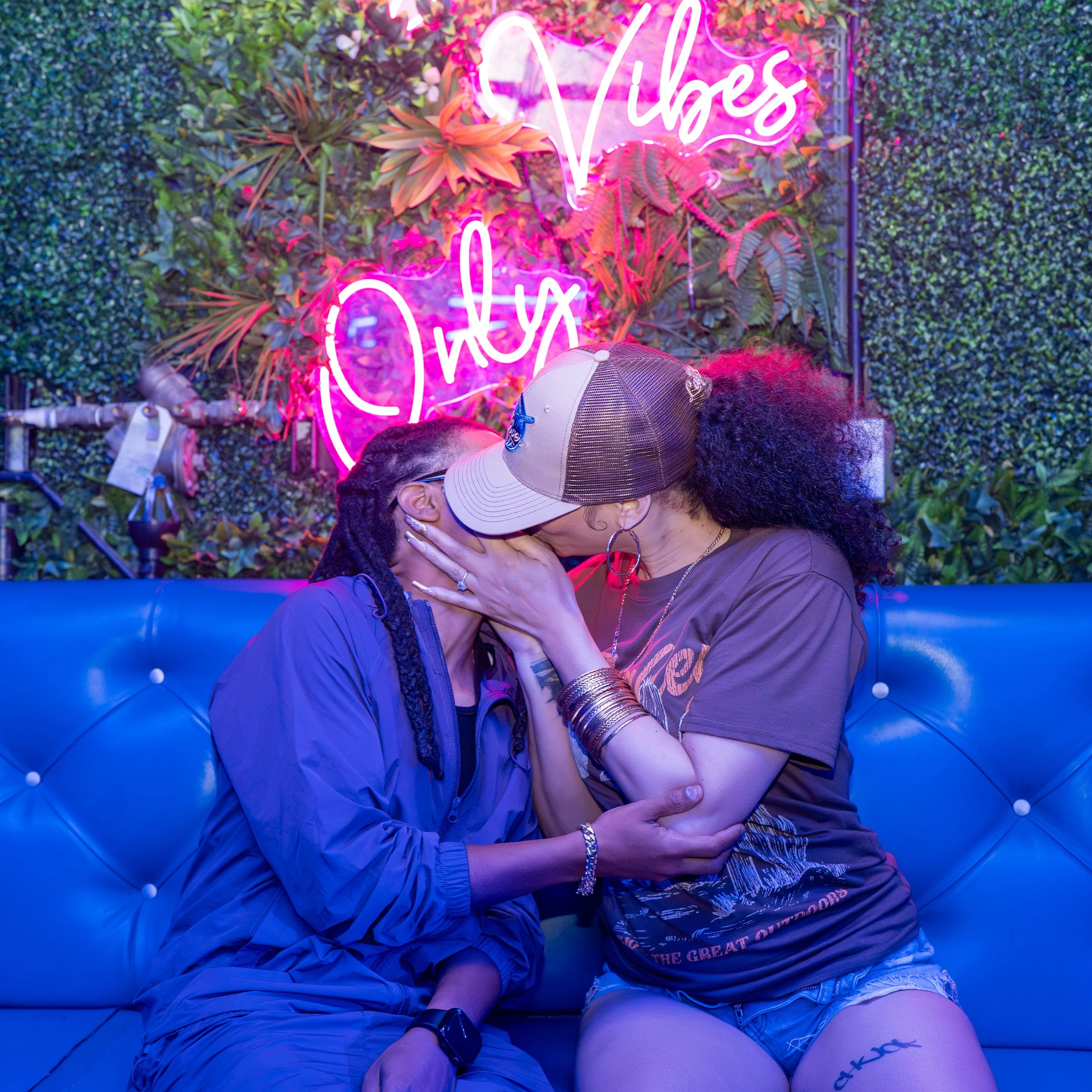 A couple kissing at a bar or lounge with a neon sign that reads 'Vibes' surrounded by plants and greenery.