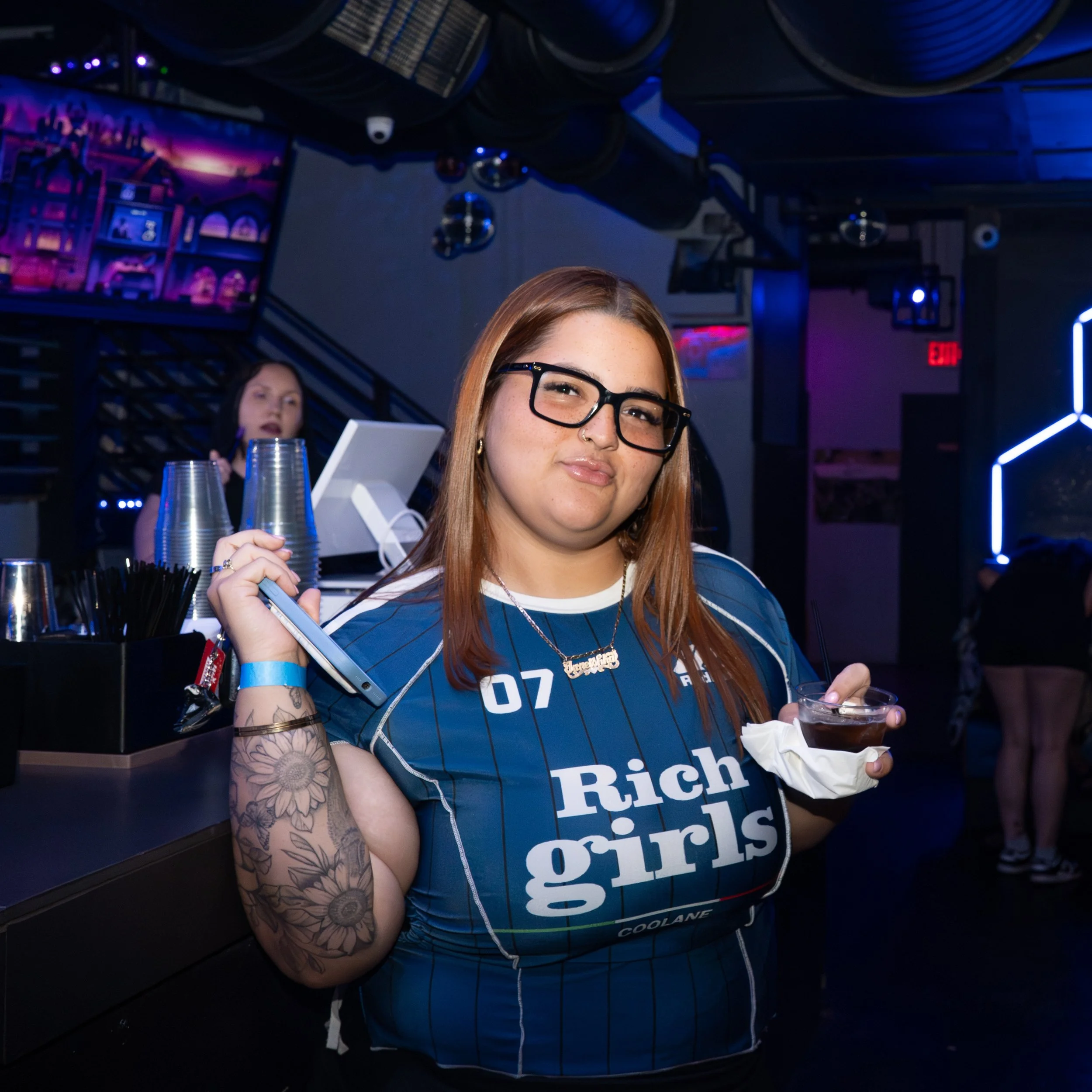 A young woman with glasses and long hair holding a drink in a plastic cup, smiling at the camera, in a dark, neon-lit bar with other patrons in the background.