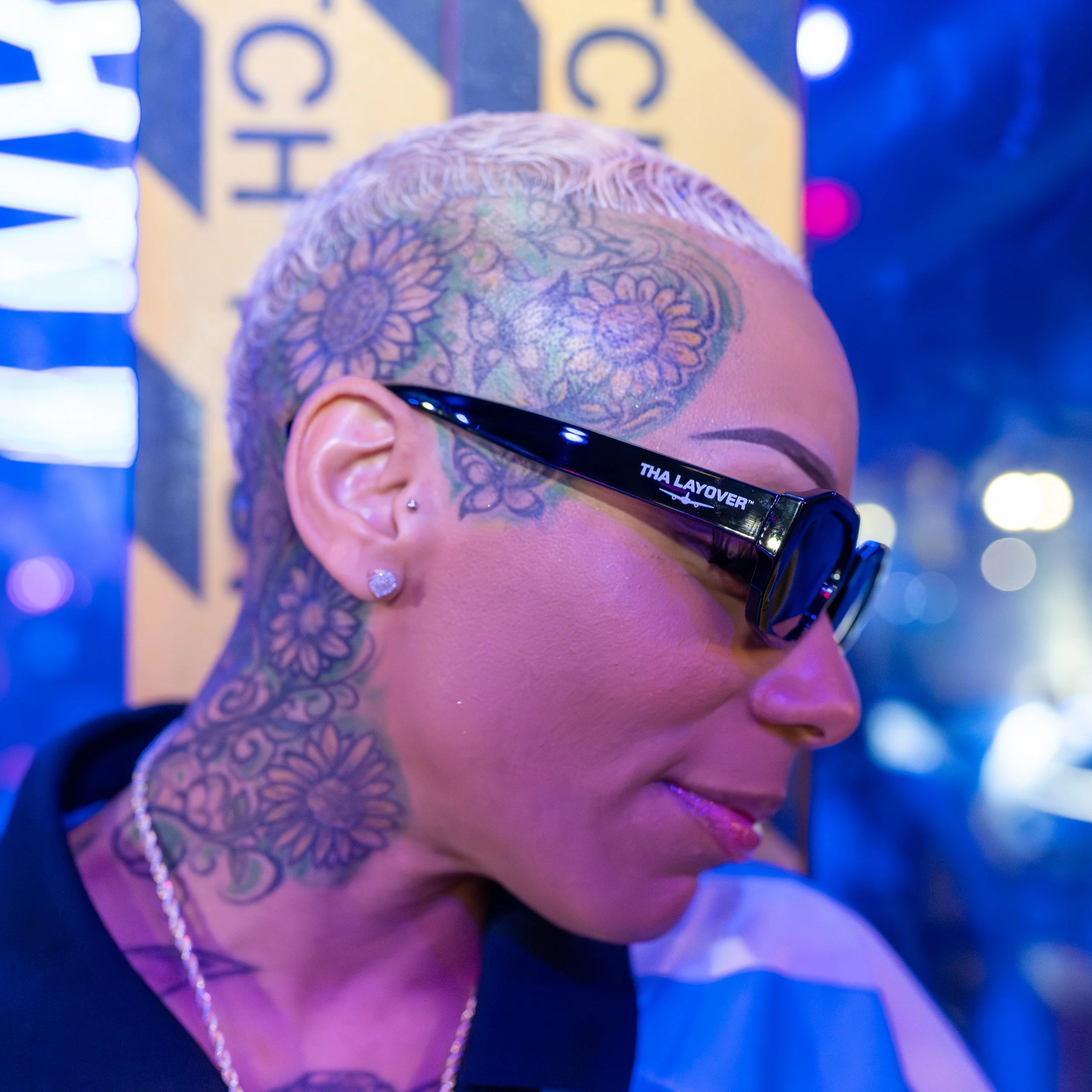 Close-up of a person's head with a tattooed scalp, wearing glasses, and a pearl necklace. The background has bright lights and a blurred sign.