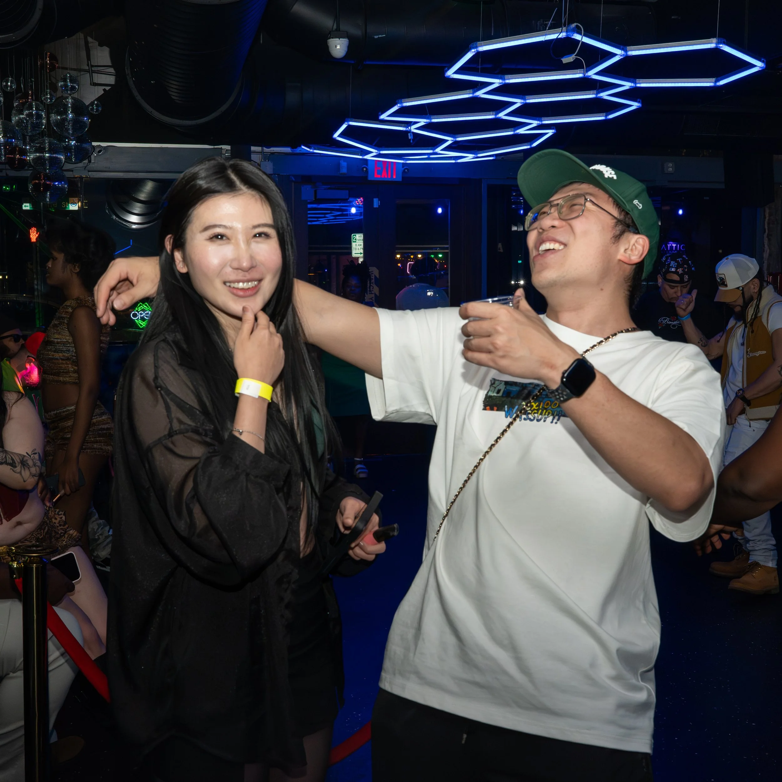 Two people smiling and enjoying drinks in a nightclub with blue neon hexagonal ceiling lights and reflective disco balls.