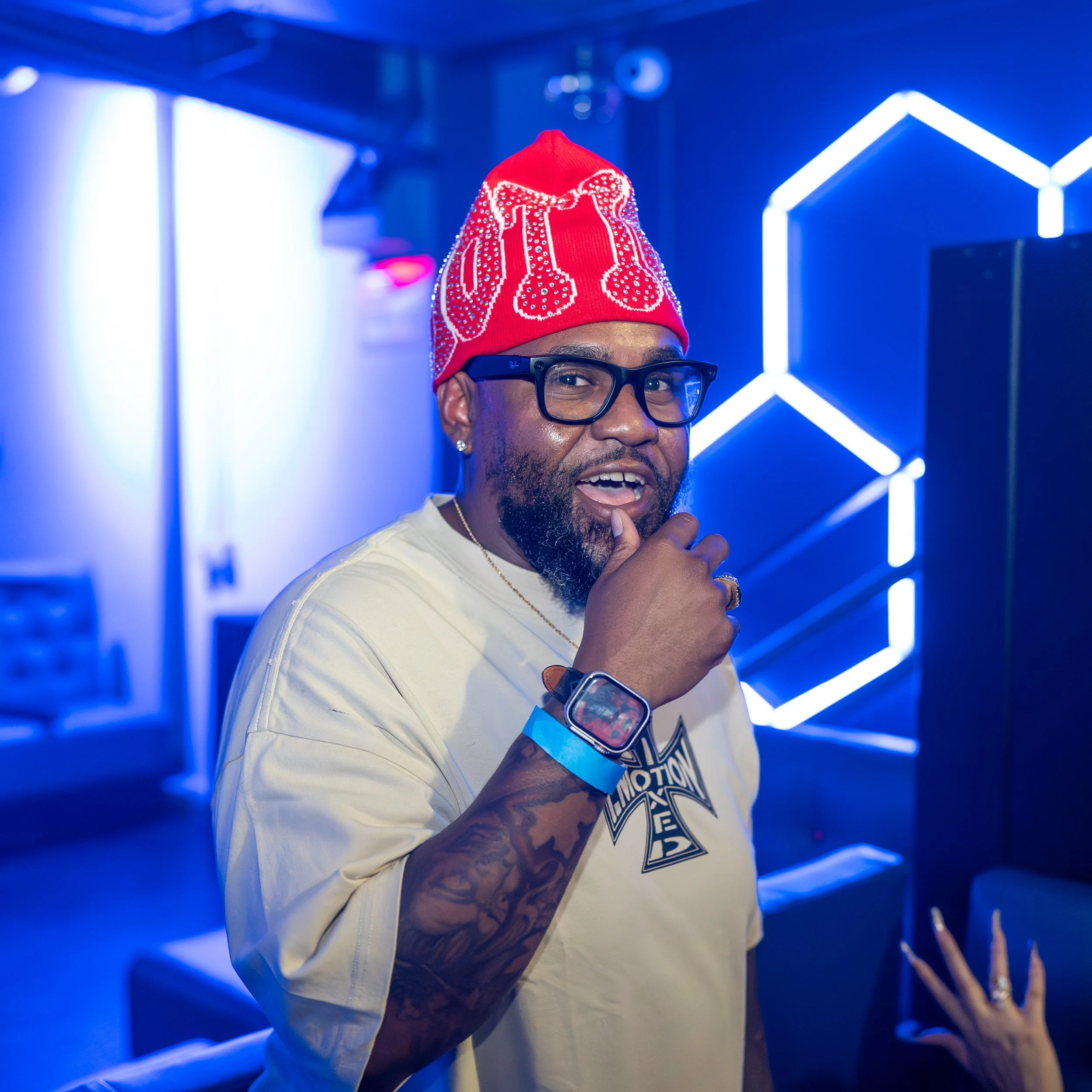 A man wearing glasses, a white T-shirt, and a red beanie with white design stands against a blue neon-lit background. He has tattoos on his left arm, a wristwatch, and is holding his chin with his right hand, smiling slightly.