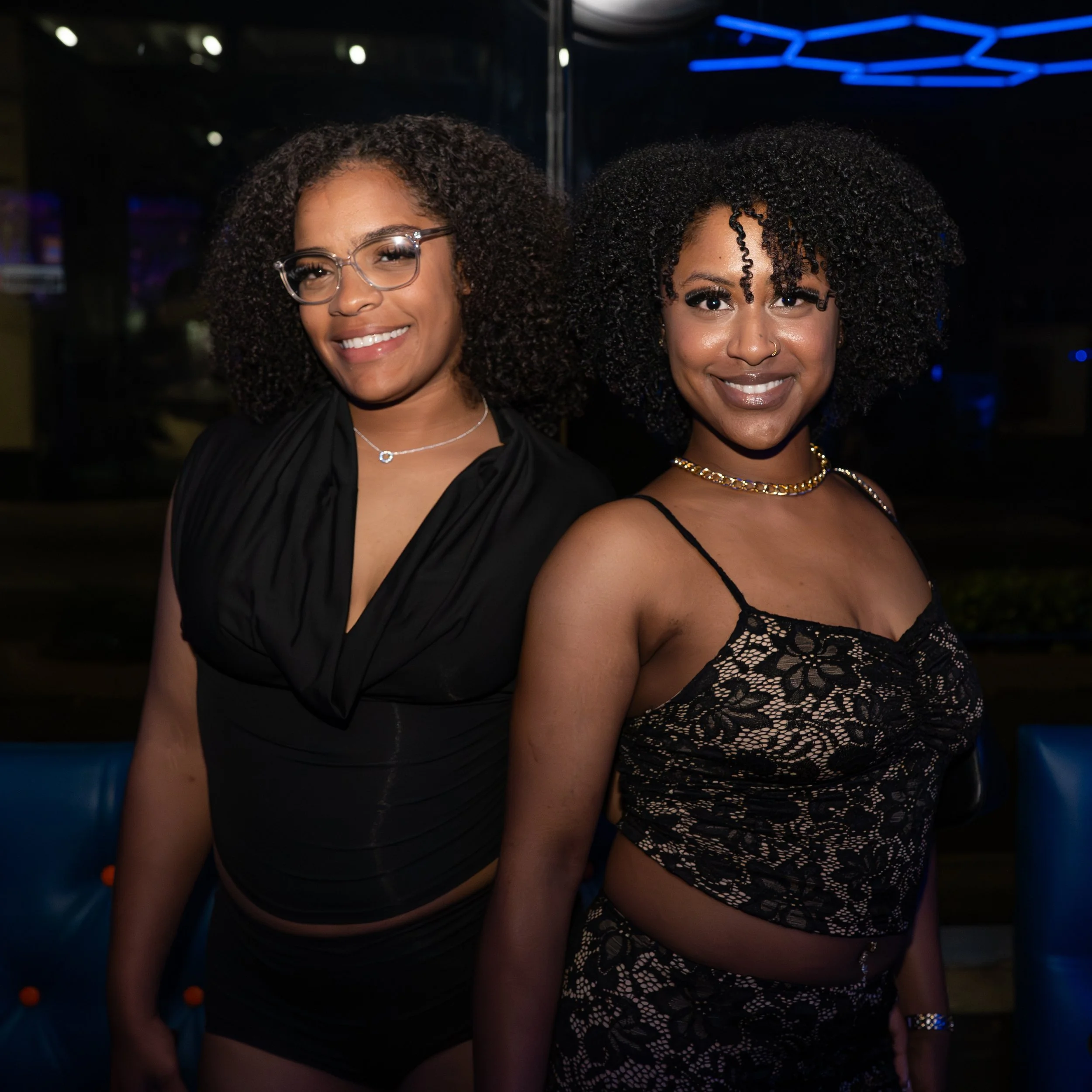 Two women with curly hair smiling in a nightlife setting, one wearing glasses and jewelry, in front of a dark background with blue neon lights.