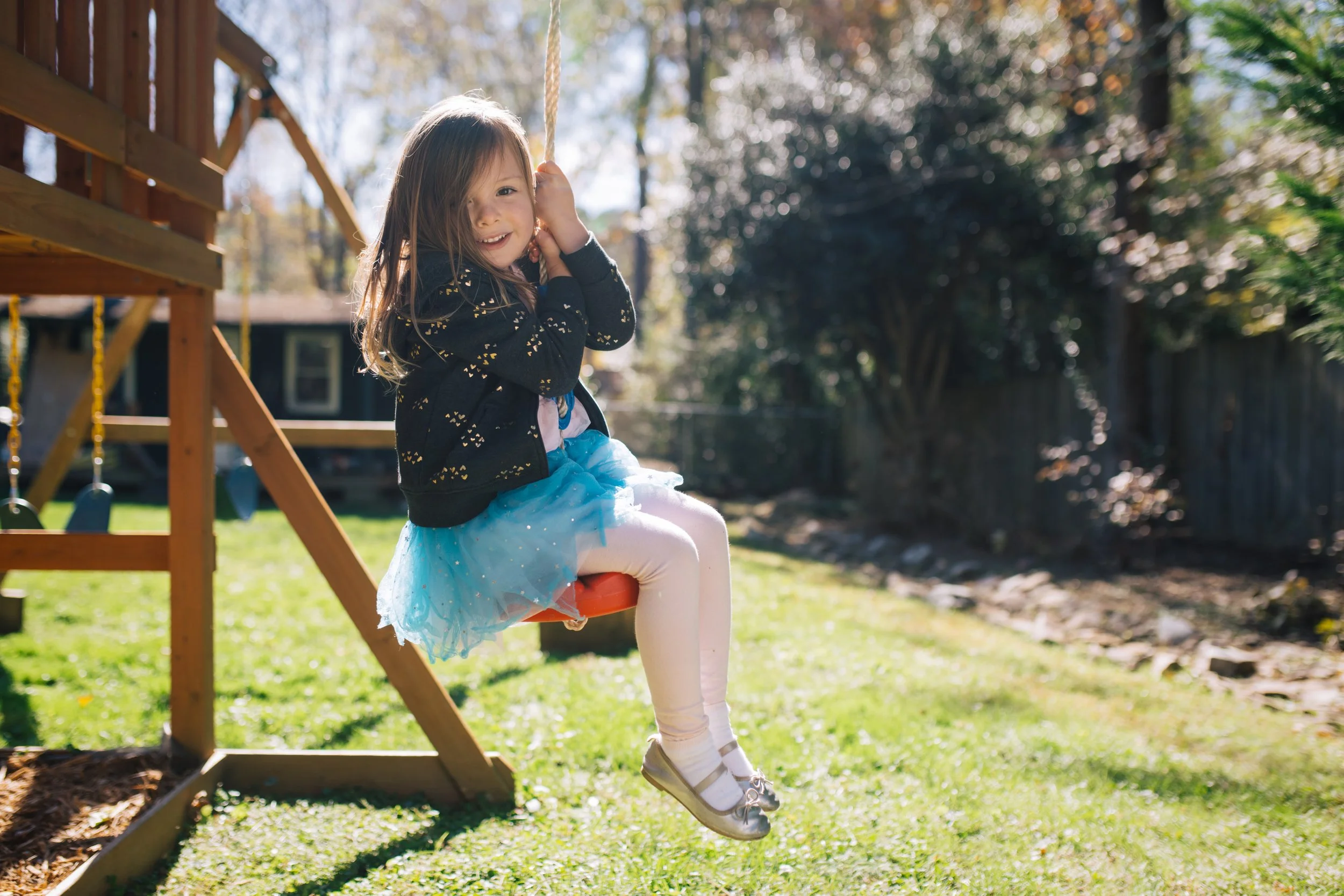 A young girl with long hair smiling while sitting on a red swing hanging from a wooden playset in a backyard during daytime, with trees and a fence in the background.