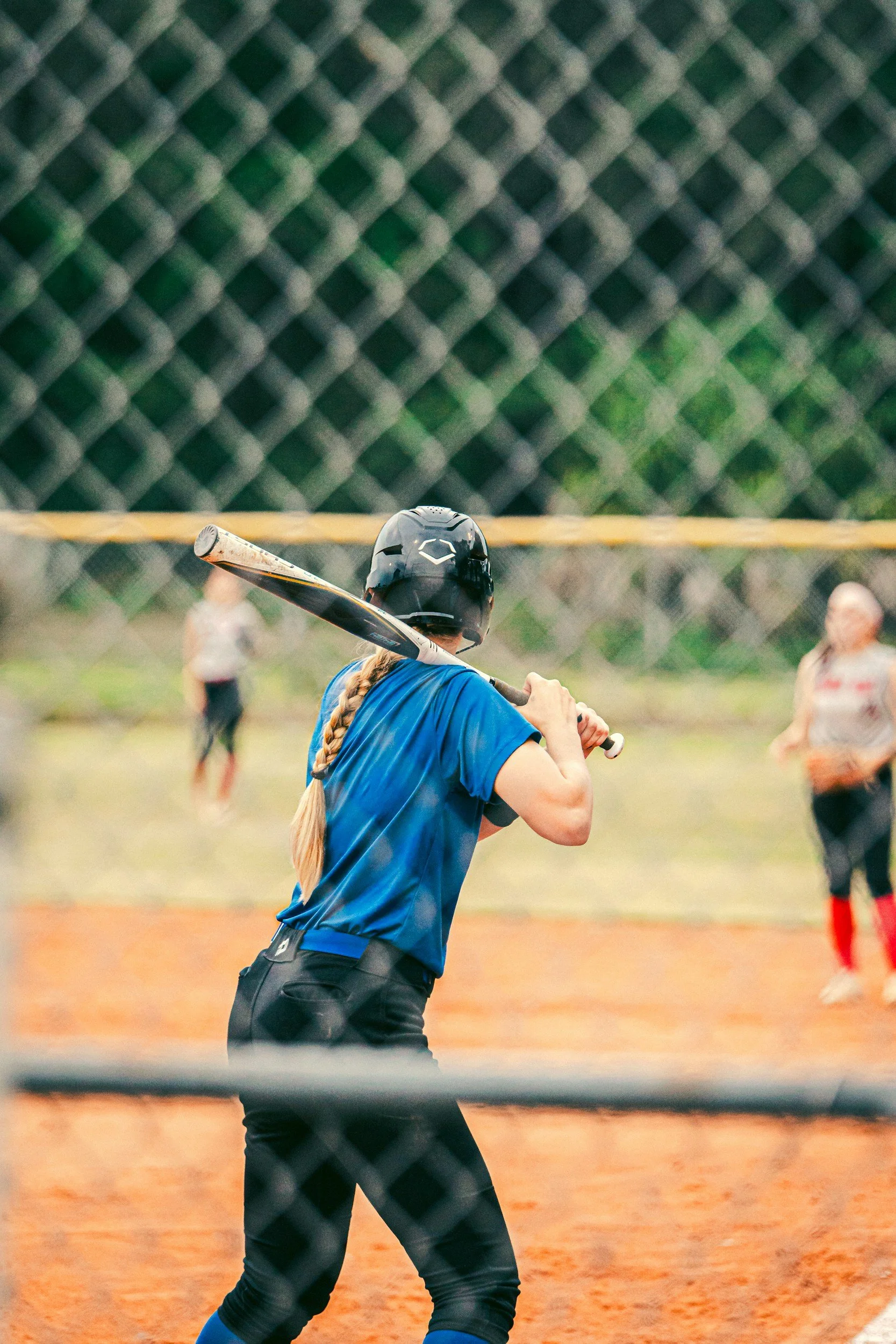 A female softball player in a blue shirt and black pants prepares to swing a bat during a game, with a chain-link fence in the foreground and other players in the background on the field.