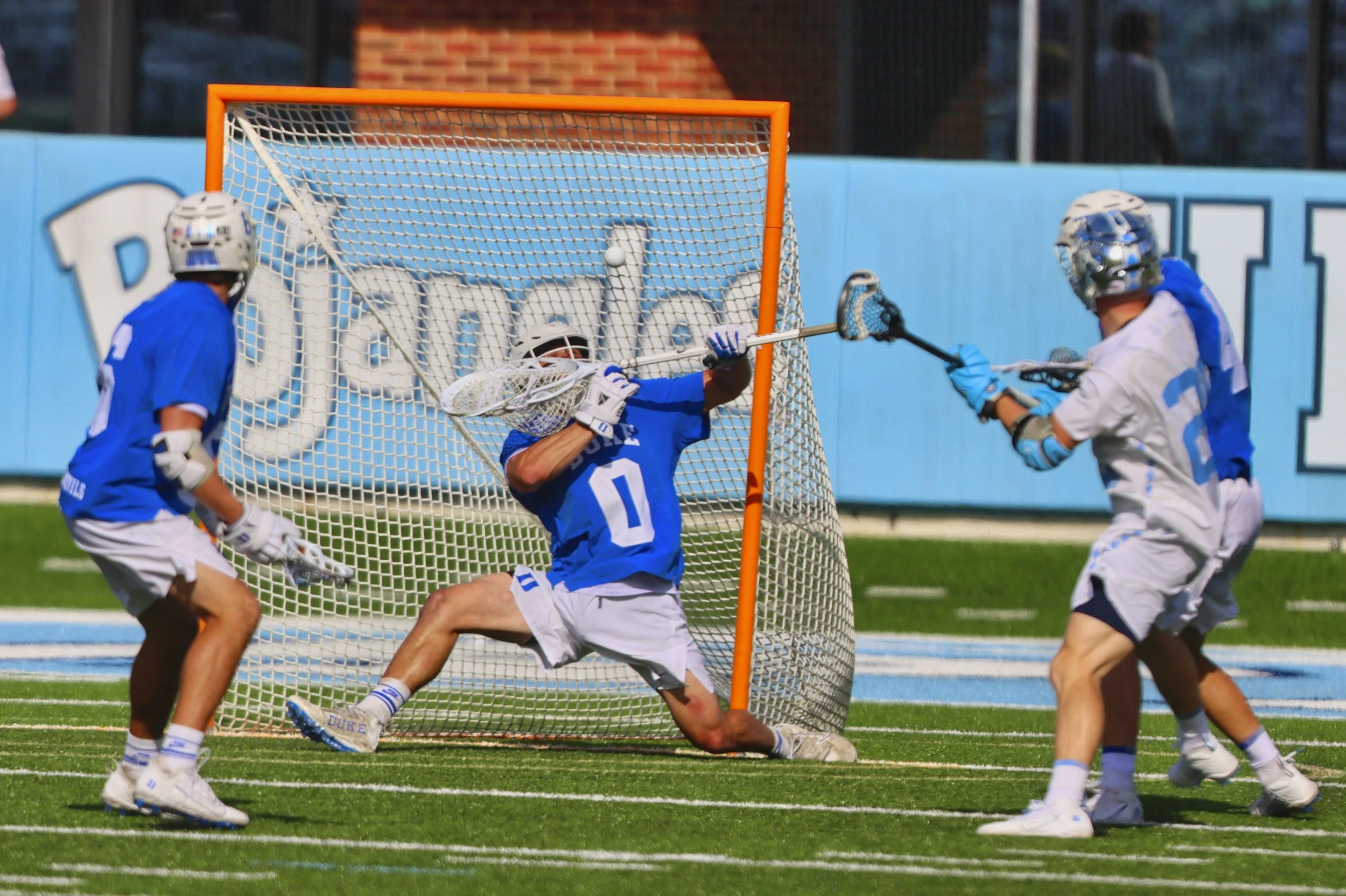 Lacrosse players in blue and white uniforms playing on a field, with one player attempting to score as the goalie and another defense player defend the net.