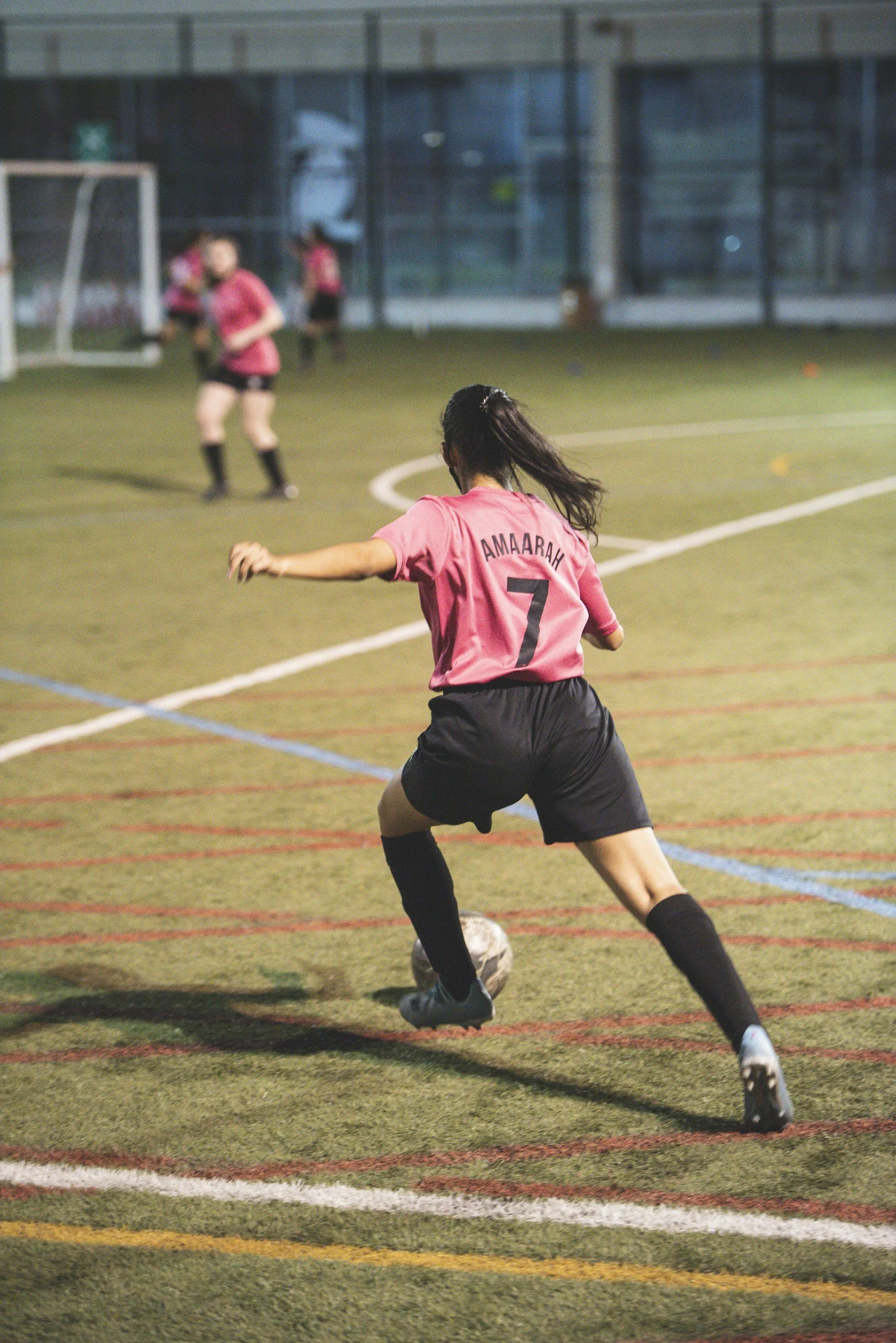 A female soccer player wearing a pink jersey with the name AMAARAH and number 7, kicking a soccer ball on a green turf field during a game or practice at night.