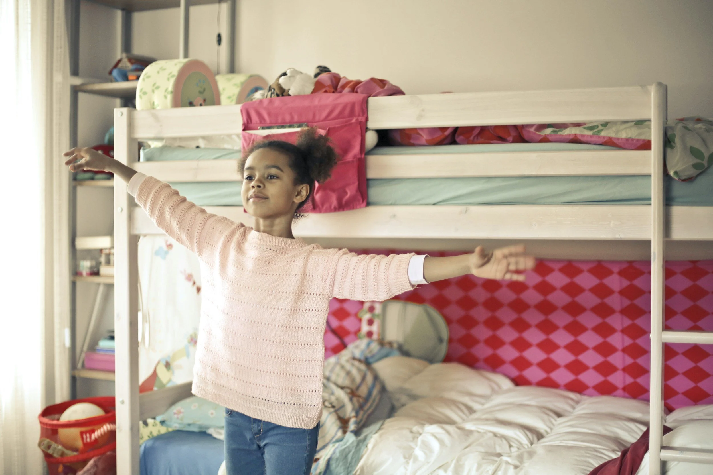 A young girl with curly hair in puffs wearing a pink sweater and blue jeans, standing in a bedroom with arms outstretched near a bunk bed, with her eyes closed and a peaceful expression.
