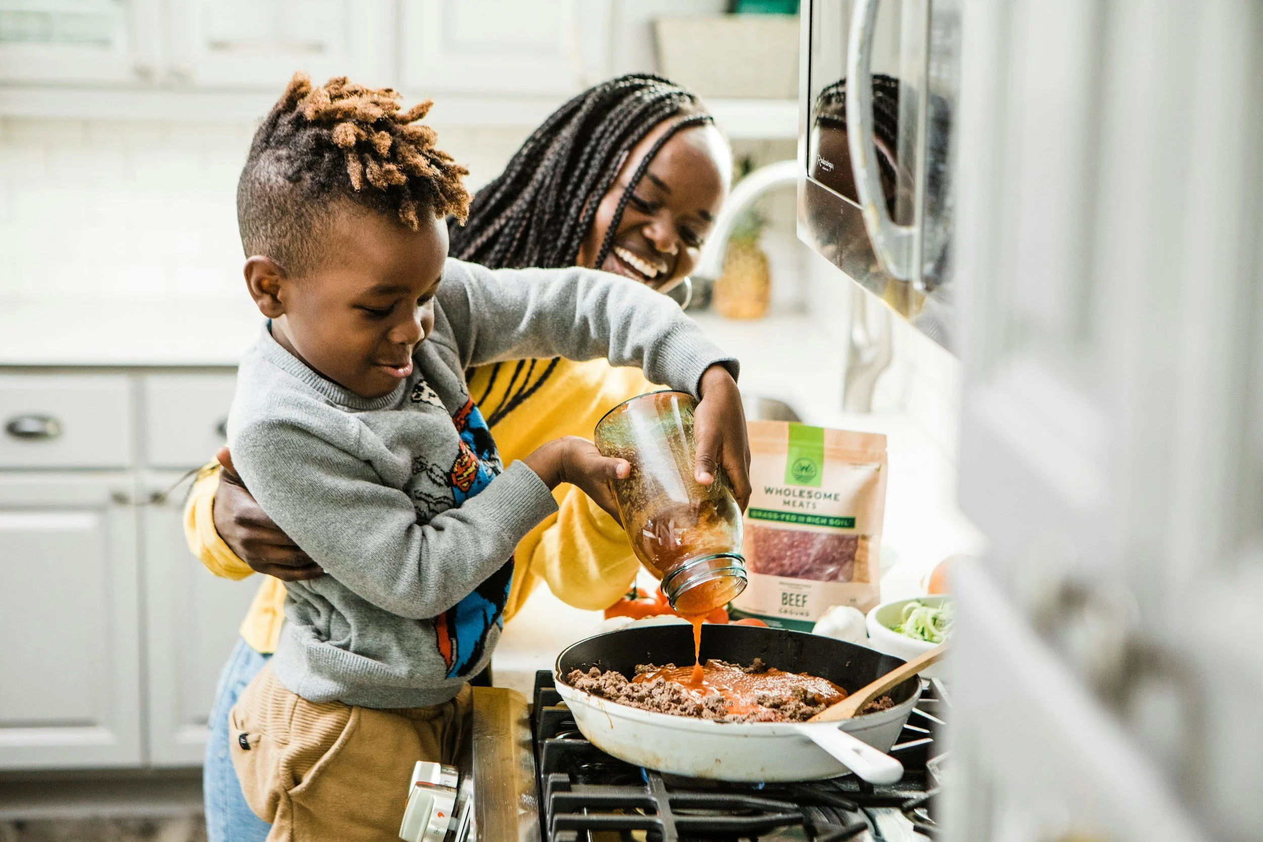 A woman and young boy cooking together in a kitchen, pouring sauce into a skillet on the stove, smiling and enjoying their activity.