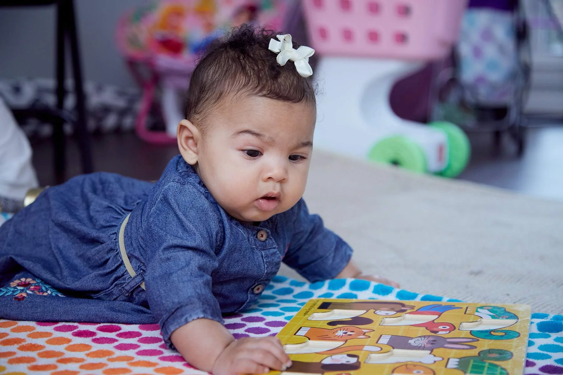 Young girl lying on her stomach on a colorful blanket, playing with a puzzle with animal pictures in a room with toys in the background.