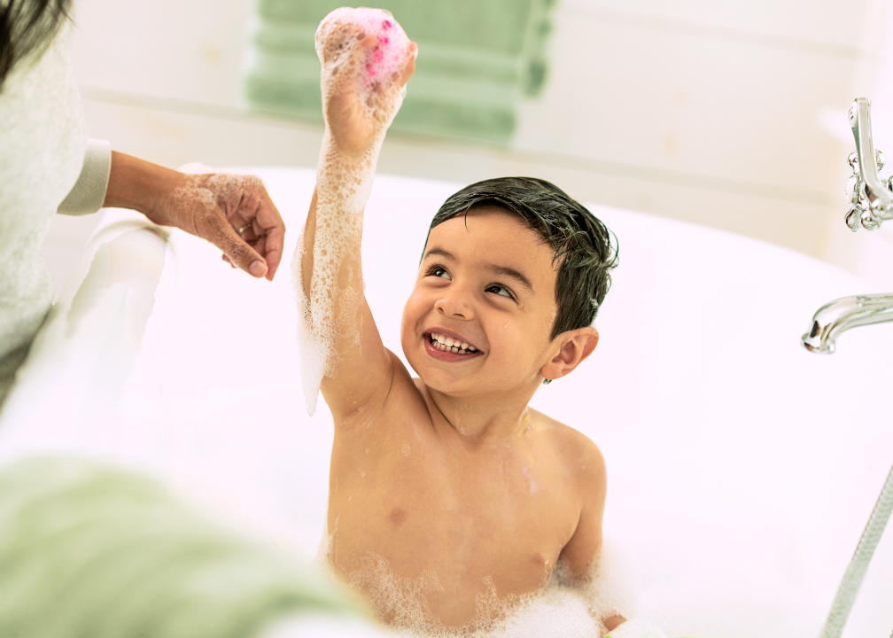 A young boy happily taking a bath in a bathtub, raising his hand with soap suds on it, while a person assist him. The boy is smiling and has wet hair.