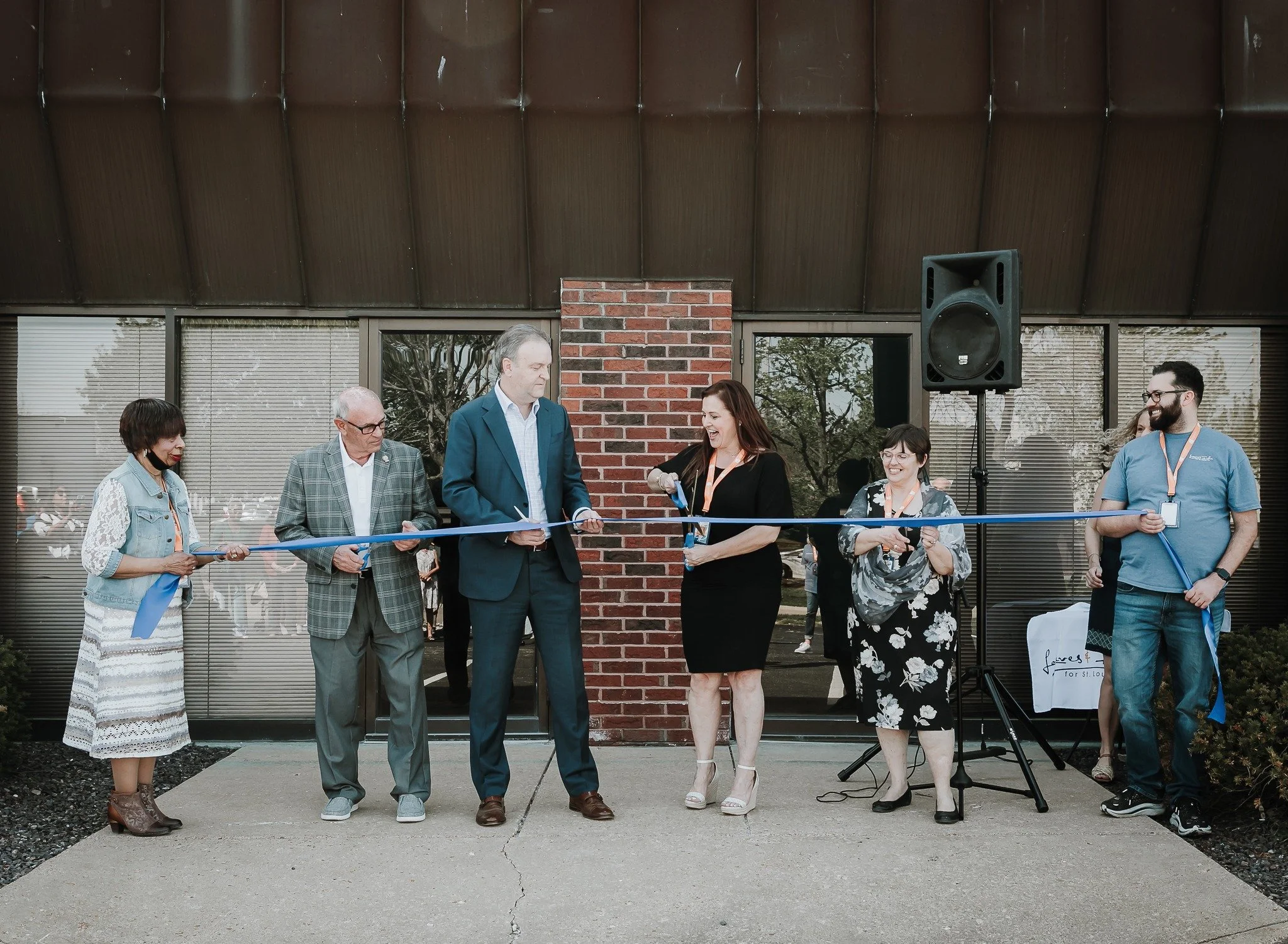 Group of people at a ribbon-cutting ceremony outside a building with brick and glass walls, with some holding a blue ribbon and scissors, celebrating an opening or event.