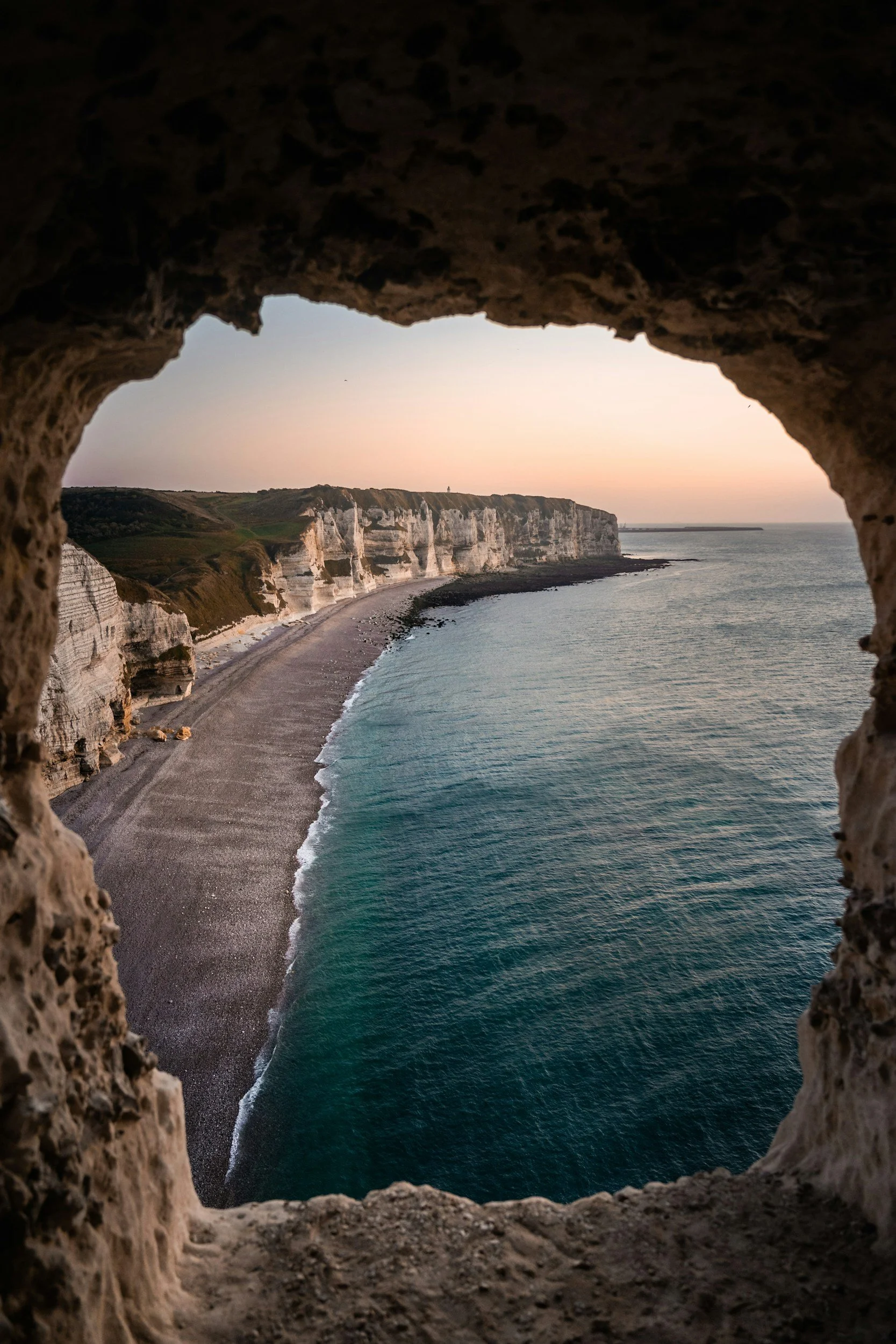 Cliffside view of a beach and the ocean, seen through a natural rock window, during sunset.