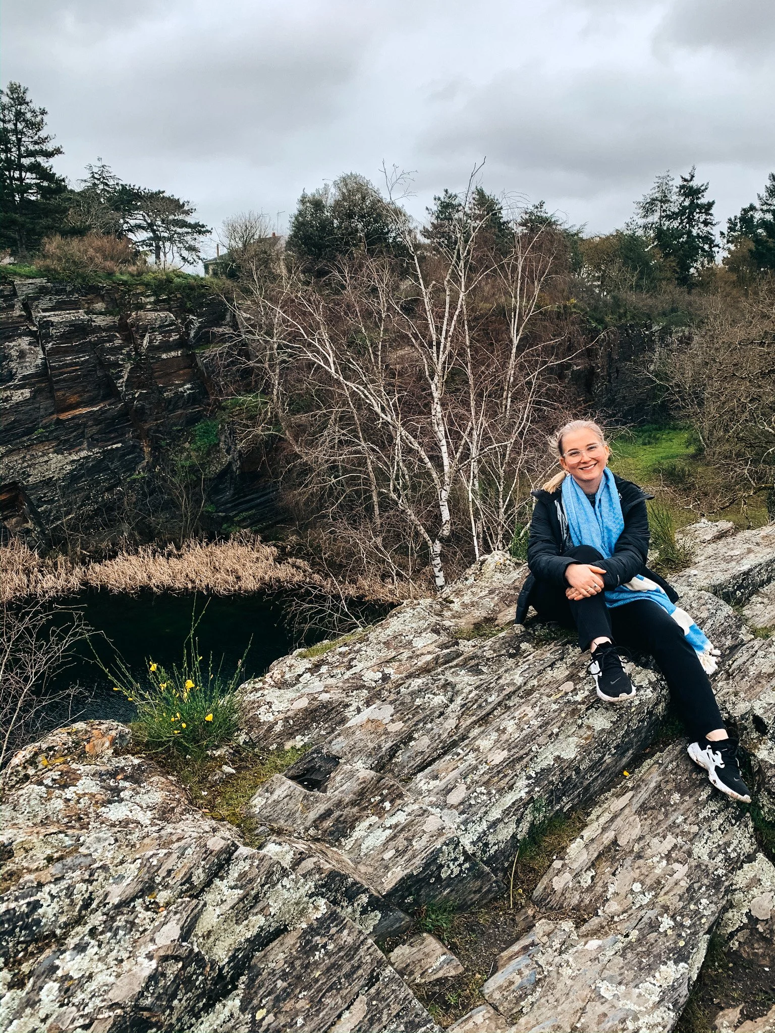 A woman sitting on a large rock outdoors with trees and a rocky landscape in the background, wearing glasses, a black jacket, black pants, black and white sneakers, and a blue scarf.