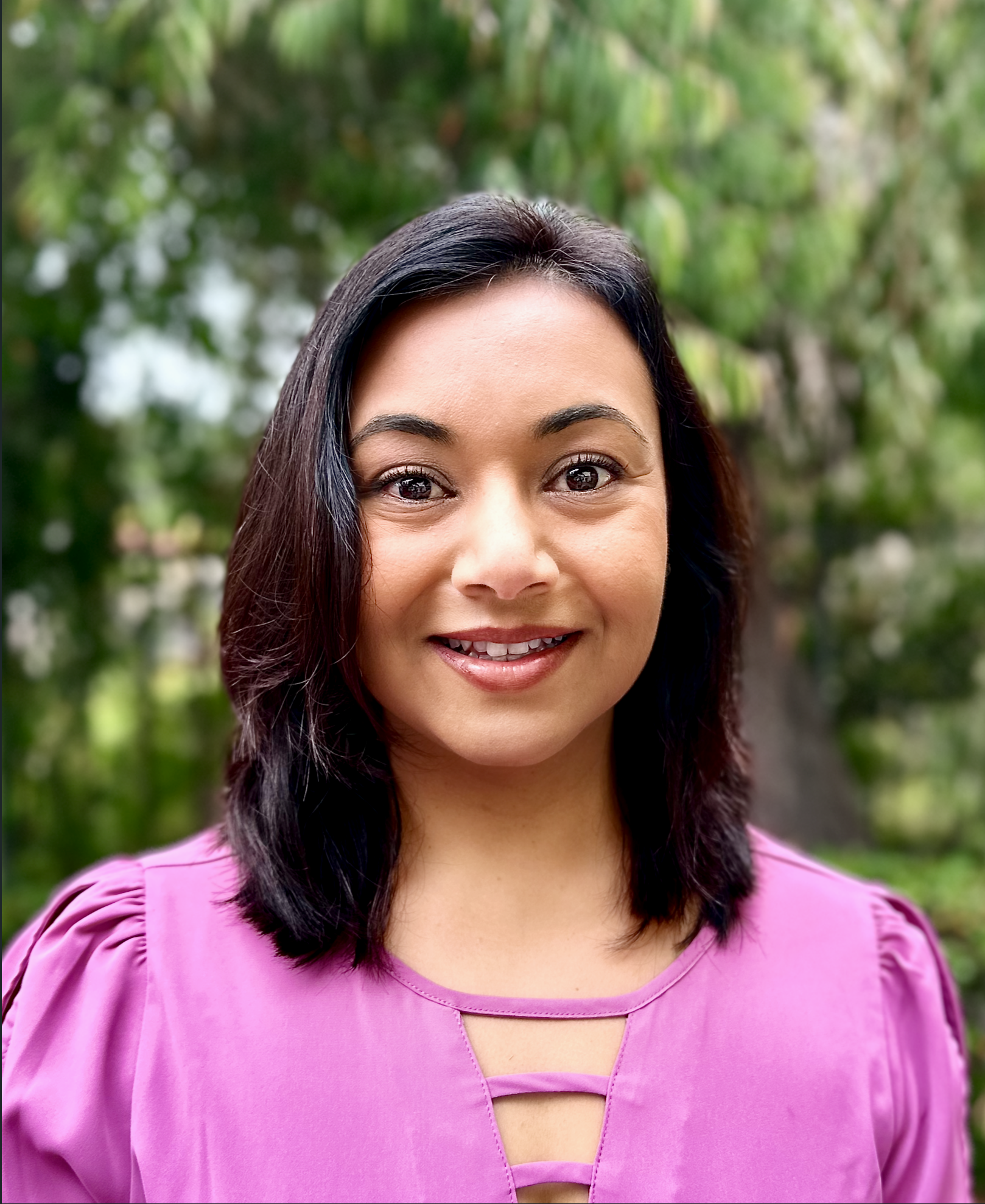 Close-up portrait of a smiling woman with shoulder-length dark hair wearing a pink top, outdoors with green foliage background.