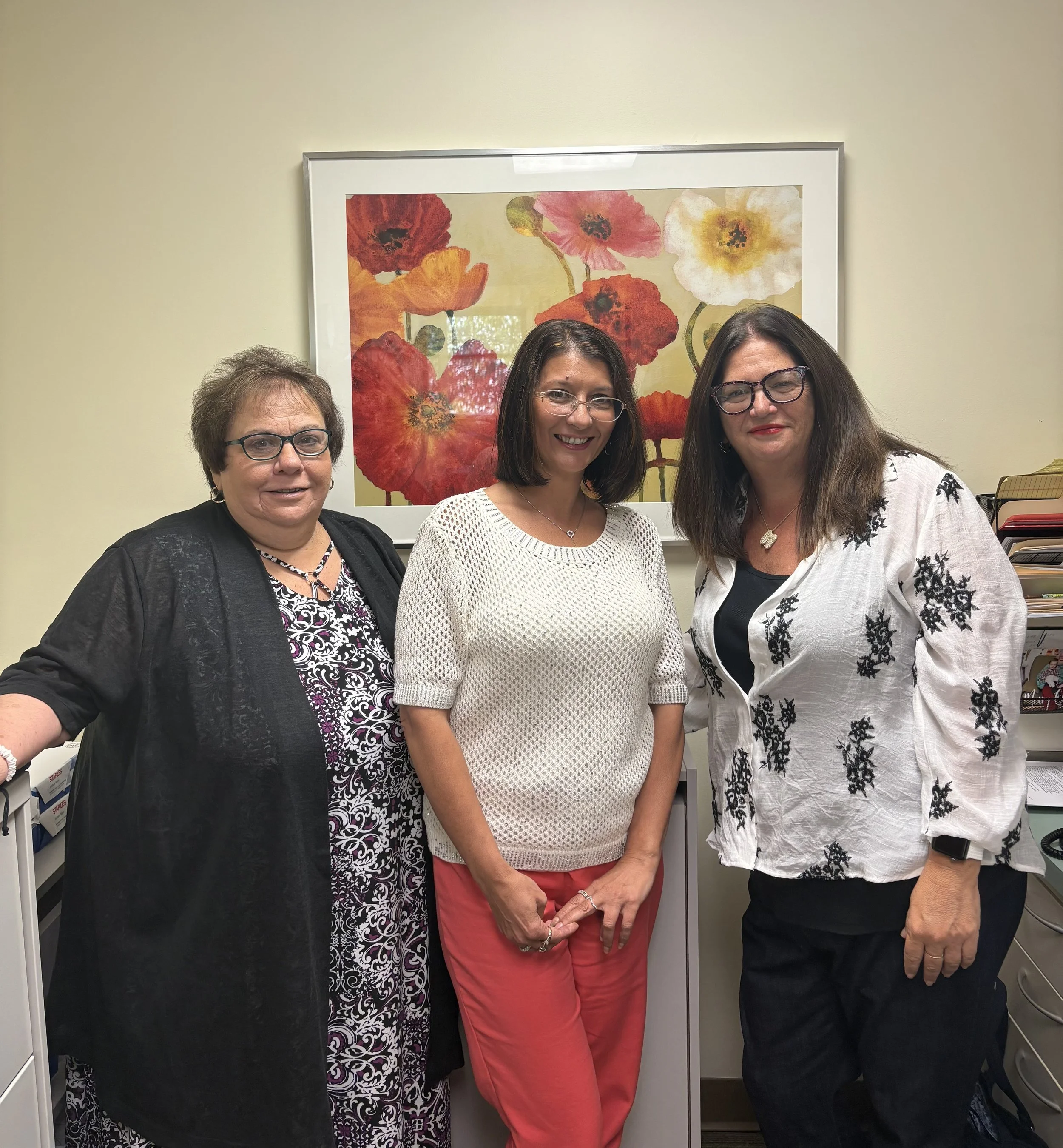 Three women standing together in an office, smiling, with a framed floral artwork behind them.