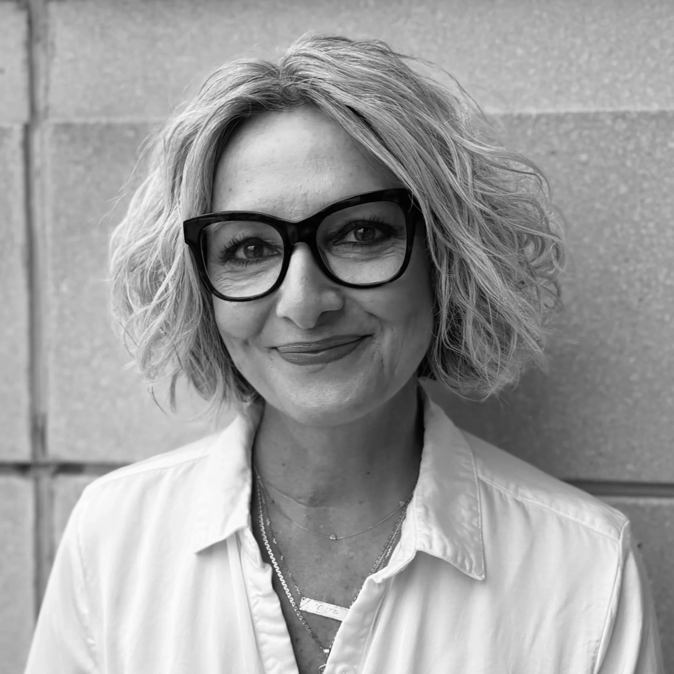 Black and white portrait of a woman with wavy hair wearing glasses, a sleeveless top, and a necklace with a cross pendant, smiling at the camera.