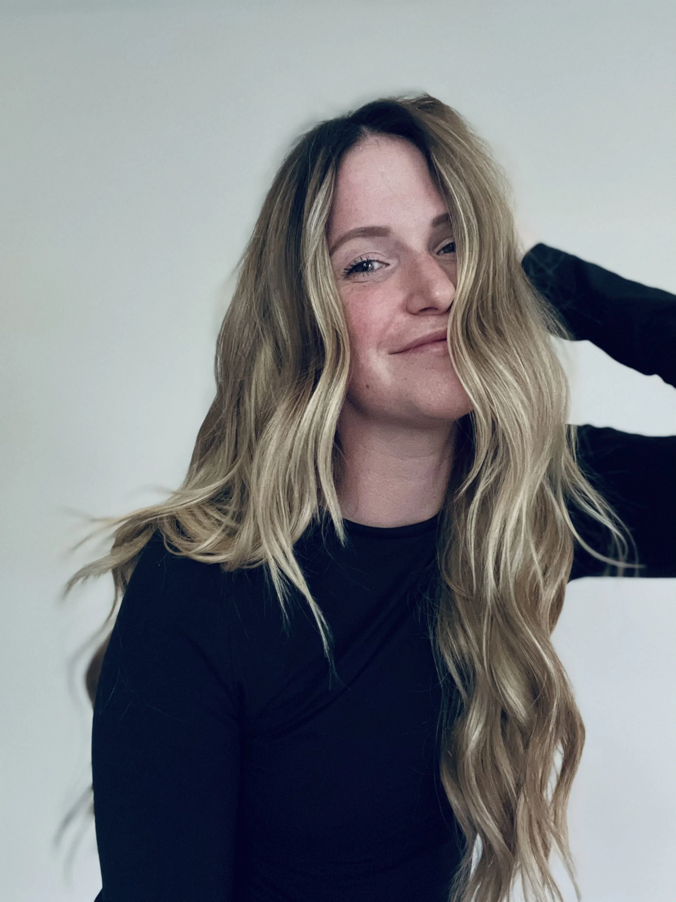 A woman with long wavy blonde hair wearing a black top, smiling with her hand behind her head, standing against a plain off-white background.