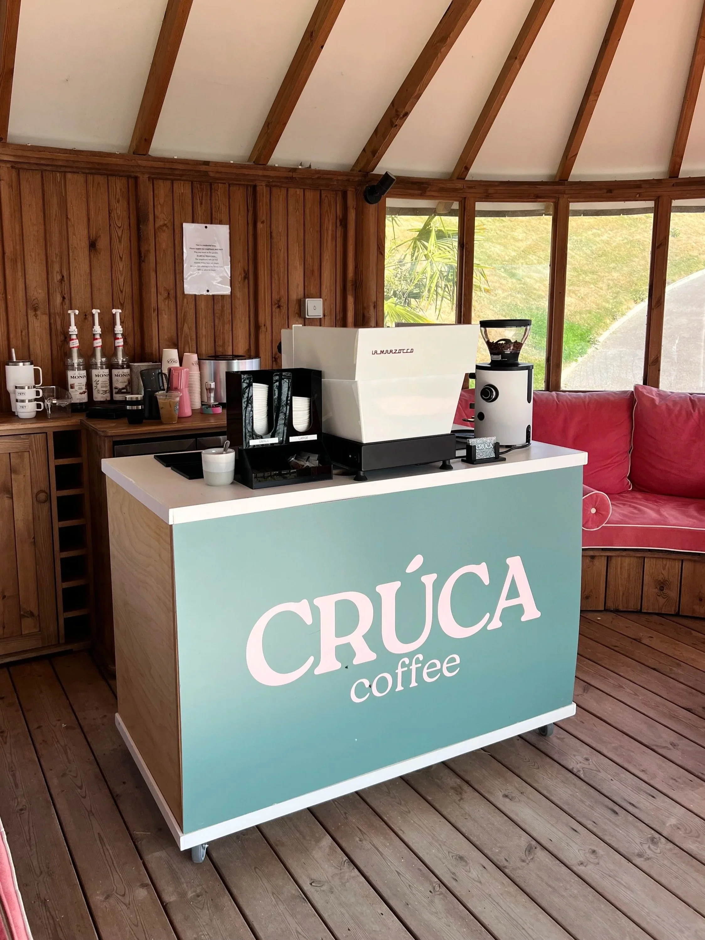 Coffee cart setup with a green counter that reads 'CRÚCA coffee', a la marzocco coffee machine, a malhkonig grinder, a white sofa, and skylights in an upstairs room.