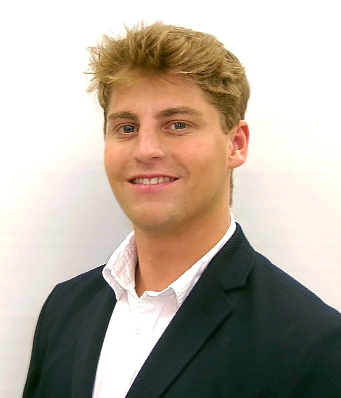 A young man with blonde hair, wearing a white collared shirt and blue suit jacket, smiling against a plain white background.